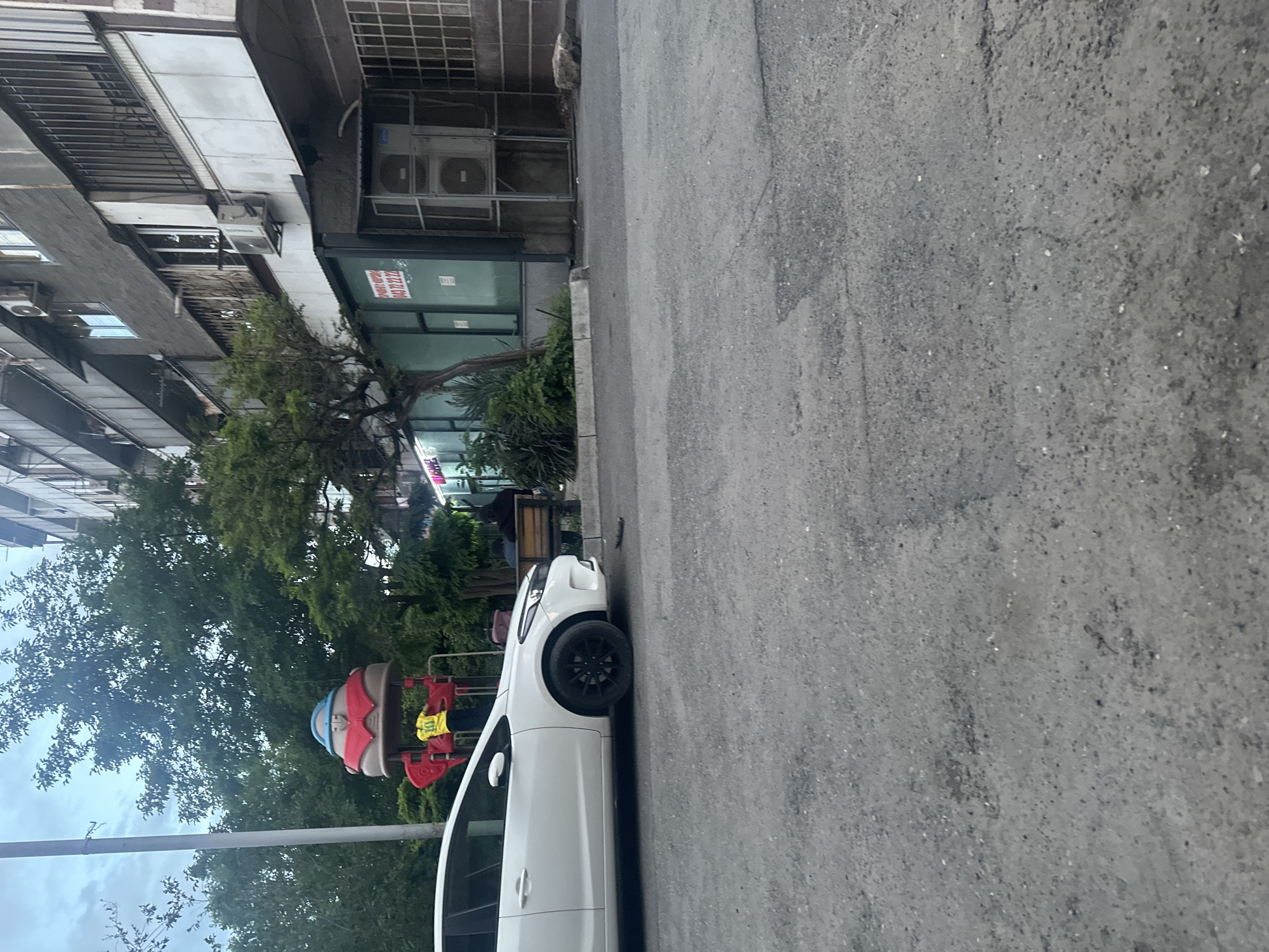 A white car is parked on the side of a street in Yerevan, Armenia. A playground with a mushroom-shaped structure is visible behind the car. The street is asphalt. A building with several floors and air conditioning units is located on the other side of the street, along with a tree and other plants. Pedestrians are visible in the background. The sky is overcast.
