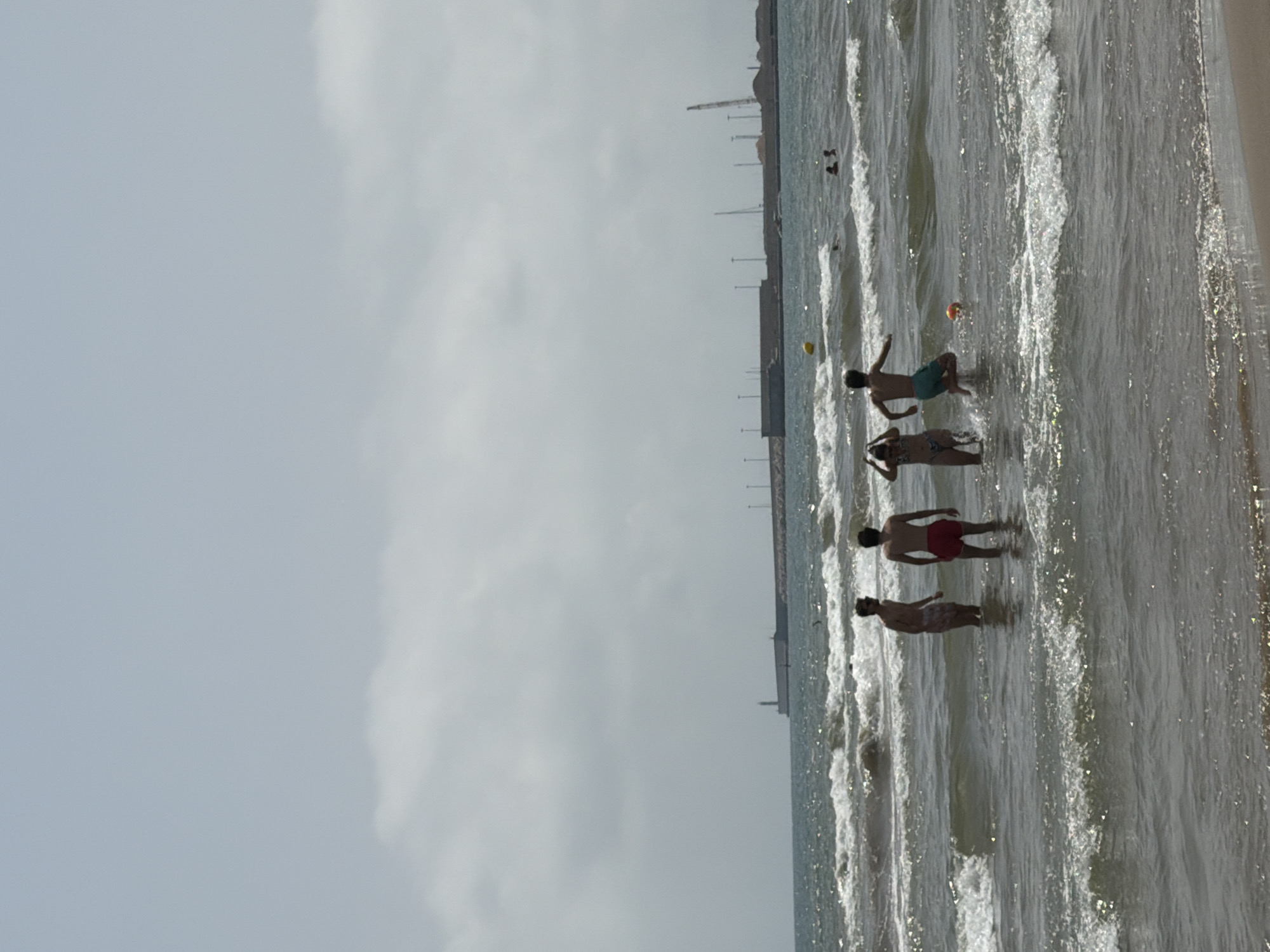 This is a media file showing a beach scene with several people in the water. The image is oriented vertically, with the ocean occupying the right side of the frame and the sky on the left.
The subjects are primarily people in swimwear, appearing to be enjoying a day at the beach. In the foreground, two individuals are visible standing in shallow water. Further back, two other individuals are also in the water, with one appearing to be reaching for a small orange object that might be a ball. In the distance, more people can be seen in the water, and what appears to be a long, dark structure, possibly a pier or a building, is visible along the coastline. There are also several poles or masts protruding from the water in the distance, suggesting a potential harbor or marina.
The activity taking place is people swimming and playing in the ocean. The water is somewhat choppy with visible waves and foam, indicating moderate conditions. The sky is overcast with diffused light, suggesting it could be a cloudy day or possibly late afternoon or early morning. The overall atmosphere seems relaxed and recreational.
Notable details include the presence of a yellow buouy and the orange object in the water. The lighting is soft due to the cloudy sky. There is no visible text in the image. The location is not explicitly identified but suggests a coastal area with some form of man-made structures in the background.
