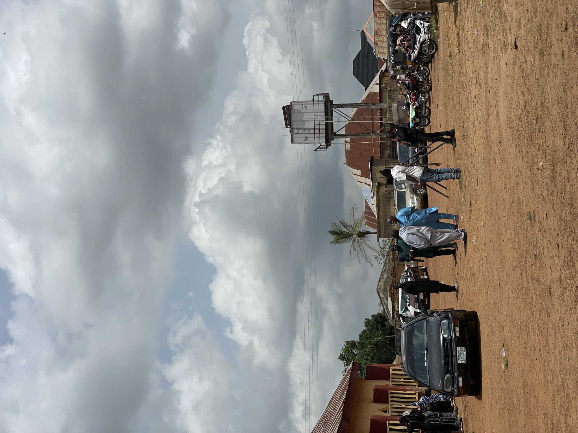 The image depicts an outdoor scene in Kassa, Nigeria, with a group of people gathered in a dirt-covered area. The sky is overcast with dramatic, dark clouds. Several buildings are visible in the background, including one with a red-tiled roof and another with a corrugated metal roof. A prominent feature is a tall, industrial-looking water tower.
In the foreground, a black Volkswagen Golf car is parked, facing the camera, with a Nigerian license plate reading "PKN 631KM." Several individuals are standing around the car and in the open area. Some are dressed in traditional Nigerian attire, while others wear casual clothing. One person is holding a camera on a tripod, suggesting a filming or photography event. Motorcycles are parked in the middle ground. The overall atmosphere appears to be a gathering or event in progress.