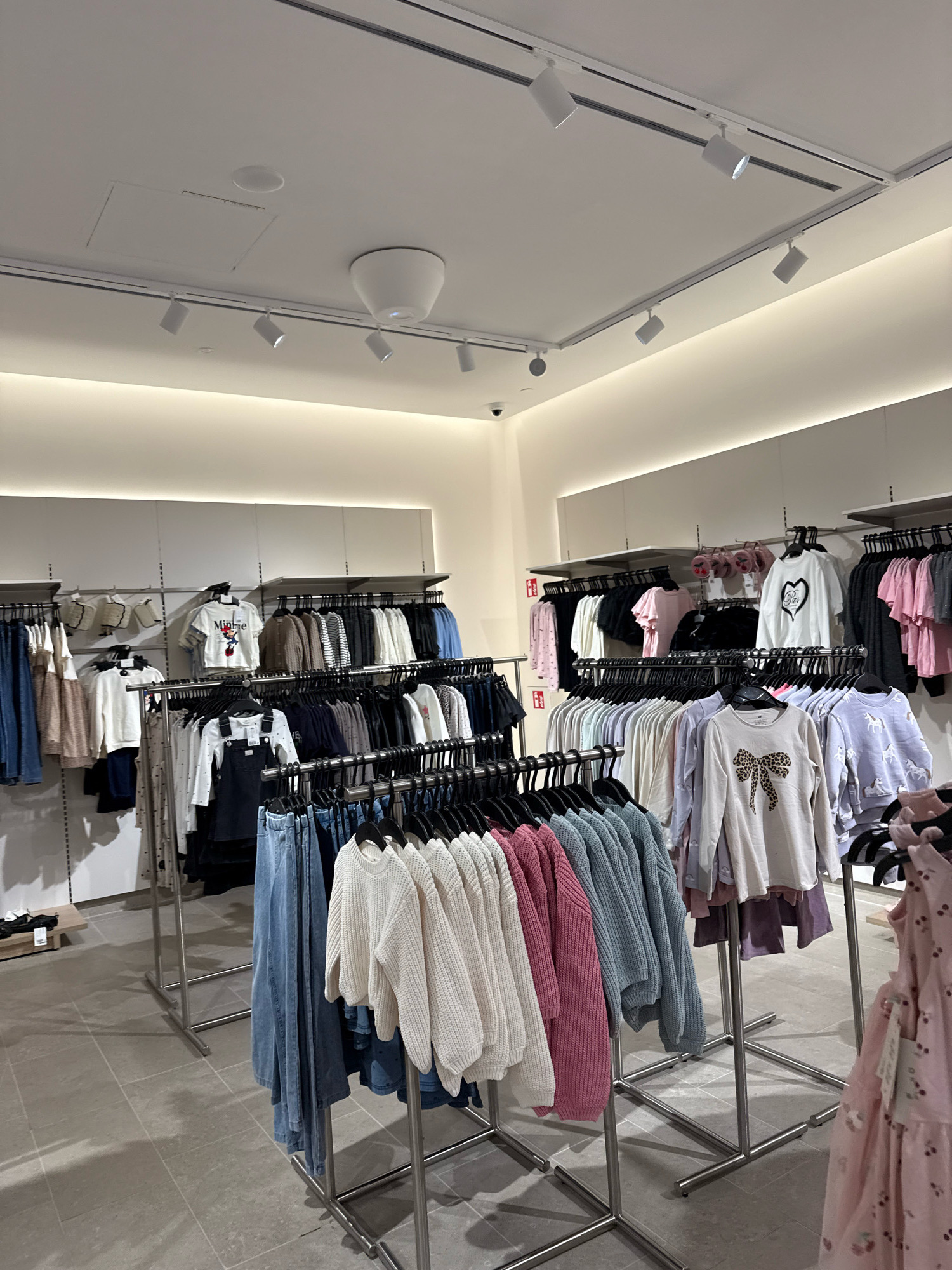A well-lit retail clothing store, likely a chain like H&M, is depicted from a medium distance, focusing on display racks of children's apparel. The store features white walls, track lighting, and a tiled floor. Numerous clothing items, including sweaters, t-shirts, dresses, and overalls, are neatly organized on metal racks. Some garments display cartoon characters like Minnie Mouse, while others feature animal prints or bows. The overall impression is one of an organized and well-stocked children's section within a modern retail environment. The image, taken indoors, does not contain any visible individuals. Based on the context of the image and potential store branding or style, it is plausible this store is located in Tallinn, Estonia, as H&M stores are present in the city.