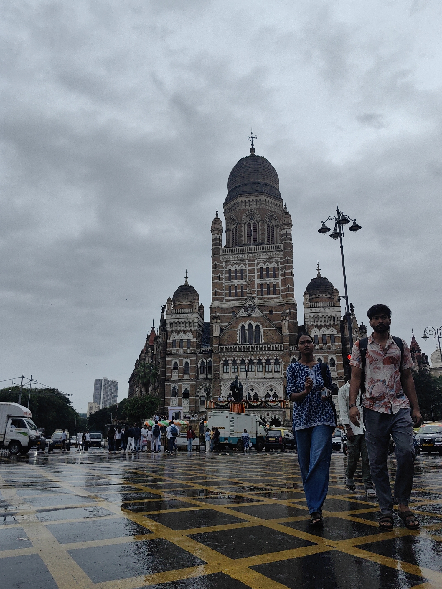 In Mumbai, India, under a heavily overcast sky, a street scene unfolds in front of the iconic Brihanmumbai Municipal Corporation building. The ground is wet and reflects the yellow crisscross markings of the road. Two individuals, a woman in a blue patterned top and jeans, and a man in a light-colored, patterned short-sleeved shirt and grey trousers, walk towards the viewer. The woman carries a dark bag over her shoulder. The man also carries a backpack. Behind them, other pedestrians are visible crossing the street, some carrying balloons. A white truck and other vehicles are present on the road. The grand Victorian Gothic architecture of the BMC building dominates the background, with its intricate details and prominent dome. The overall atmosphere suggests a cloudy day, possibly after rain.