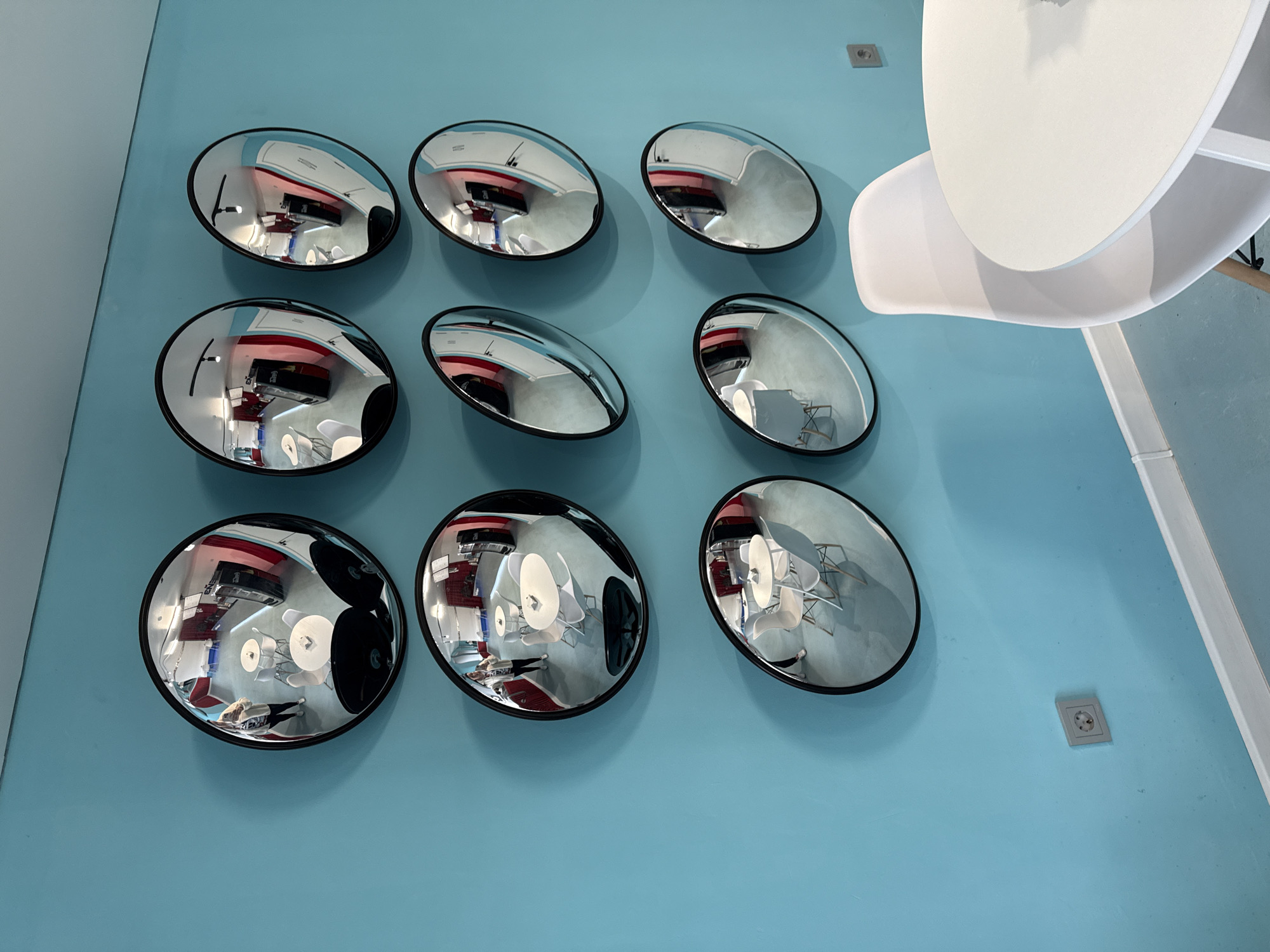 The image depicts a light blue wall on which nine circular convex mirrors are mounted in a 3x3 grid. Each mirror reflects a distorted image of an interior space, including tables, chairs, and possibly a person. A white table and chair partially occupy the upper right of the frame. Two electrical outlets are visible on the wall. The location appears to be in Chaikovsky, Russia, based on the architectural and interior design elements reflected in the mirrors, which align with common styles in that region.