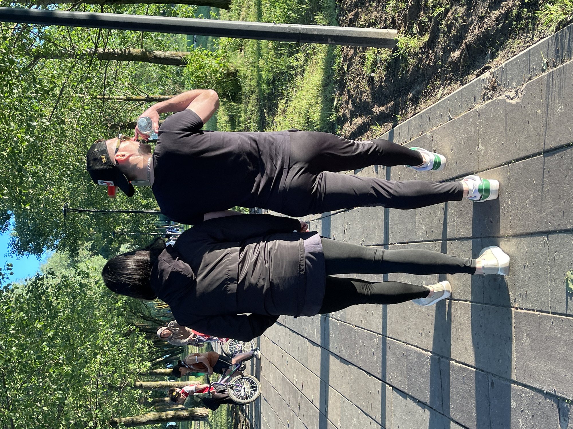 In a sunlit park, two individuals, seen from behind, walk along a paved pathway lined with trees. The person on the left wears a black hooded jacket, dark leggings, and white sneakers. To their right, a person in a black t-shirt, black pants, and black baseball cap drinks from a clear plastic water bottle. Their footwear consists of white sneakers with green accents. In the background, other park-goers are visible, including a person on a bicycle and others walking amongst the trees. The environment suggests a warm, clear day.