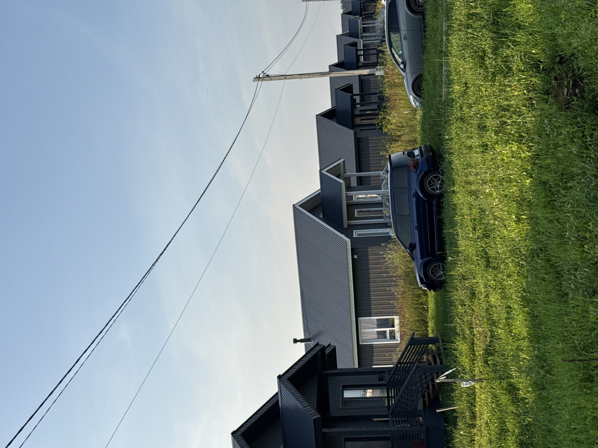 The image depicts a residential setting with several modern, dark-colored houses lined up along a grassy area. Two cars are visible: a dark blue minivan parked on the grass and a grey sedan parked further back. The houses feature corrugated metal roofs and large windows. In the background, more houses and trees can be seen. A wooden utility pole with several wires stretches across the sky, which is a clear light blue, suggesting it is daytime, likely late afternoon or early evening due to the warm, soft lighting. The overall scene is peaceful and quiet, typical of a suburban or rural environment. There is no visible text in the image. The location context suggests this is in Podshivalovo, Russia.