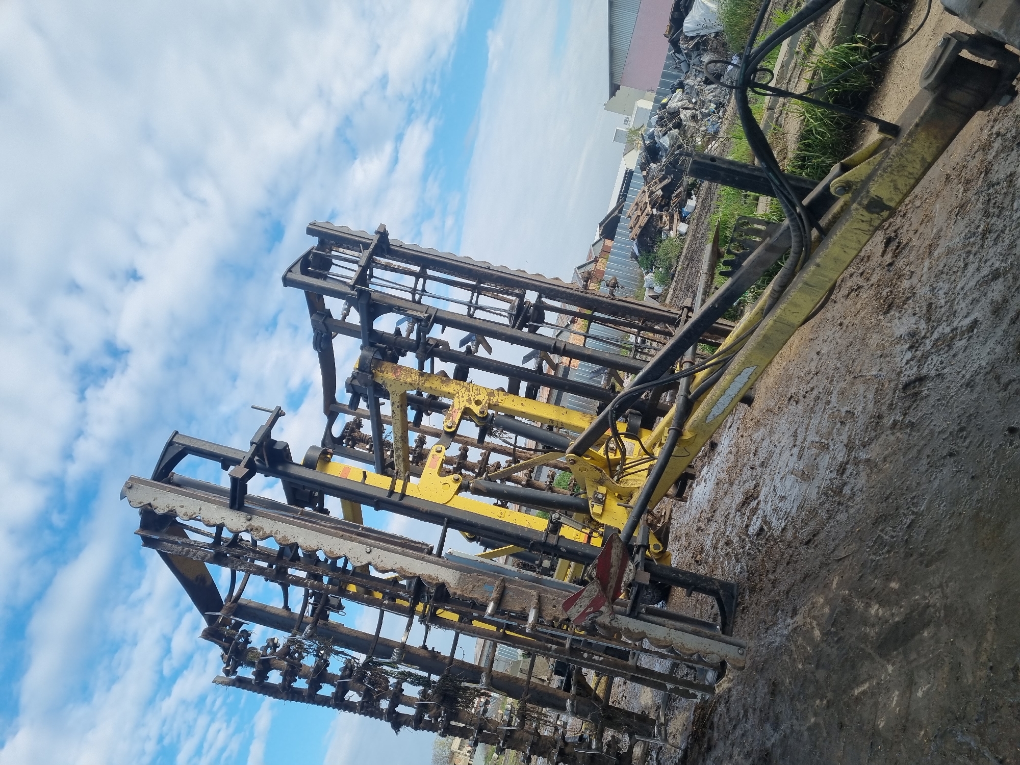 The image shows a piece of agricultural machinery, likely a cultivator or harrow, sitting on muddy ground. The machinery is composed of a heavy metal frame with multiple rows of tines or teeth designed to break up soil. The frame is painted in a combination of black and yellow. The surrounding environment appears to be a storage area or yard, possibly in an industrial or agricultural area. In the background, there is a pile of scrap metal and what seems to be a building with a corrugated metal roof. In the background a blue sky with white clouds is visible. The location is possibly Ovcharovo, Bulgaria, based on the prompt. No people are visible in the image.
