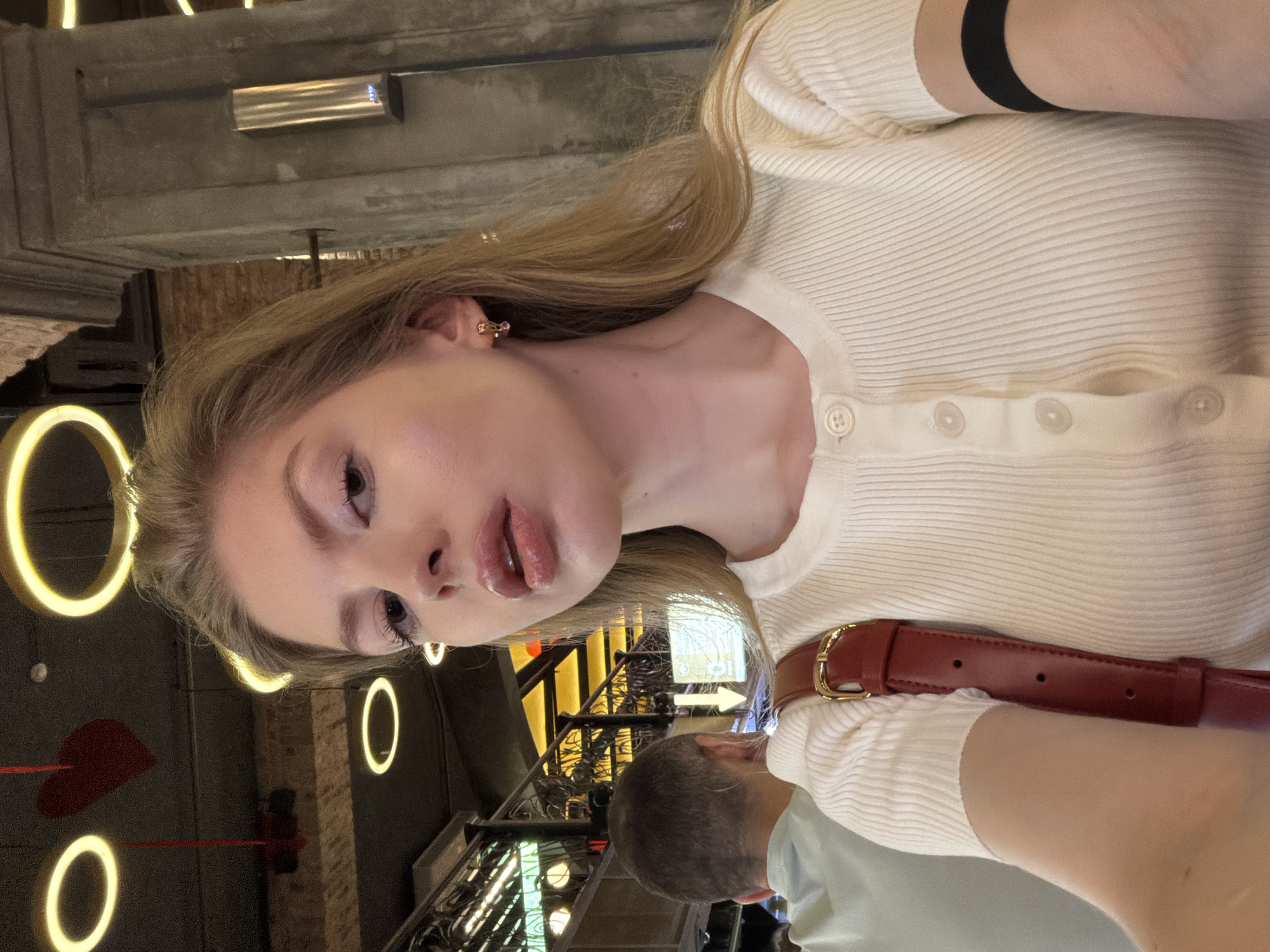A young woman with blonde hair, wearing a white ribbed top with buttons, is taking a selfie. She is wearing earrings and has a black band on her arm. A red leather purse strap is visible. A man with gray hair is partially in the frame below her. The background is blurred, but appears to be a restaurant or bar with decorative lighting, including circular lights and a red heart-shaped decoration. It's possibly a location in Moscow, Russia. The precise location cannot be determined with certainty from the image.