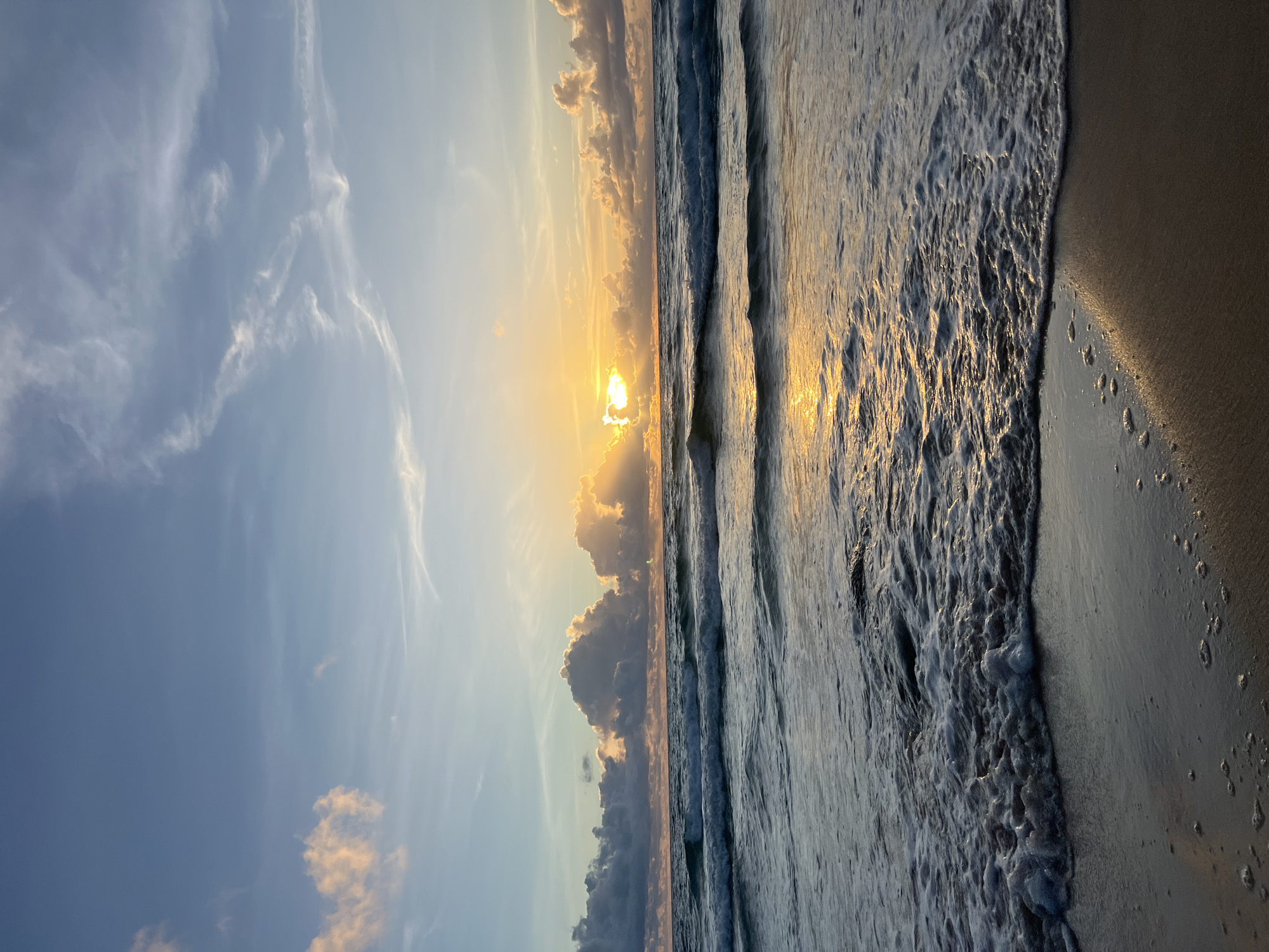 An ocean waves crash against a sandy shore at sunrise in Choeng Thale, Thailand. The sky is a gradient of soft blues and oranges, with wispy clouds illuminated by the rising sun peeking through. The sun's rays cast a warm glow on the water, creating a shimmering reflection. The waves are gentle, with white foam washing onto the wet sand. The overall scene is serene and peaceful, capturing the beauty of a tropical morning.