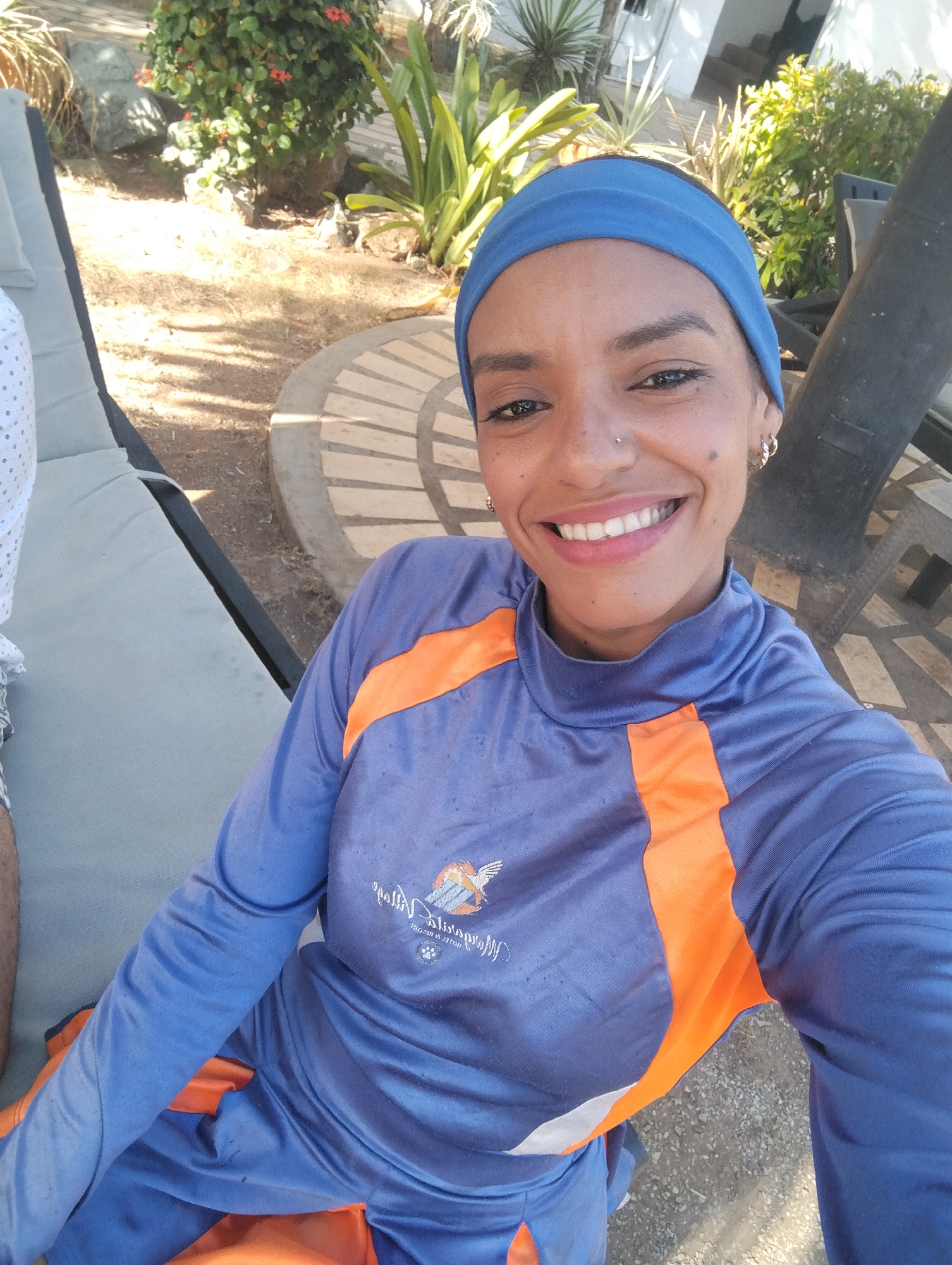A woman with a blue headband smiles directly at the camera. She wears a blue and orange long-sleeved top with a logo on the chest. She has pierced ears and a nose ring. She is sitting on a gray cushioned lounge chair outdoors, with greenery and a light-colored building in the background. The location is likely a resort in the city of Porlamar, Venezuela, based on the style of the surroundings.