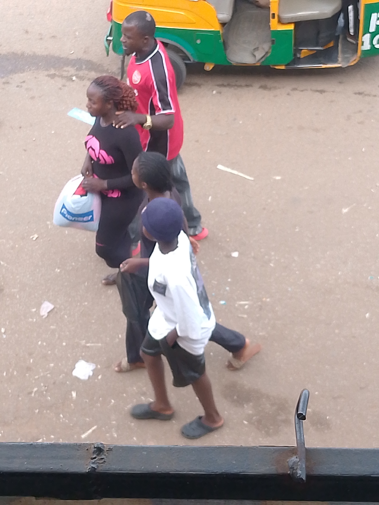 In Bukuru, Nigeria, a man in a red and black shirt with a gold watch on his wrist is seen walking with a woman wearing a black top with pink floral patterns, carrying a white and blue "Pioneer" branded bag. Behind them, a younger person in a white t-shirt and dark shorts, wearing a blue cap, walks with another individual in dark clothing. They are on a paved surface, likely a road, with a yellow and green auto-rickshaw parked in the background. The scene appears to be during the daytime, with muted lighting conditions.