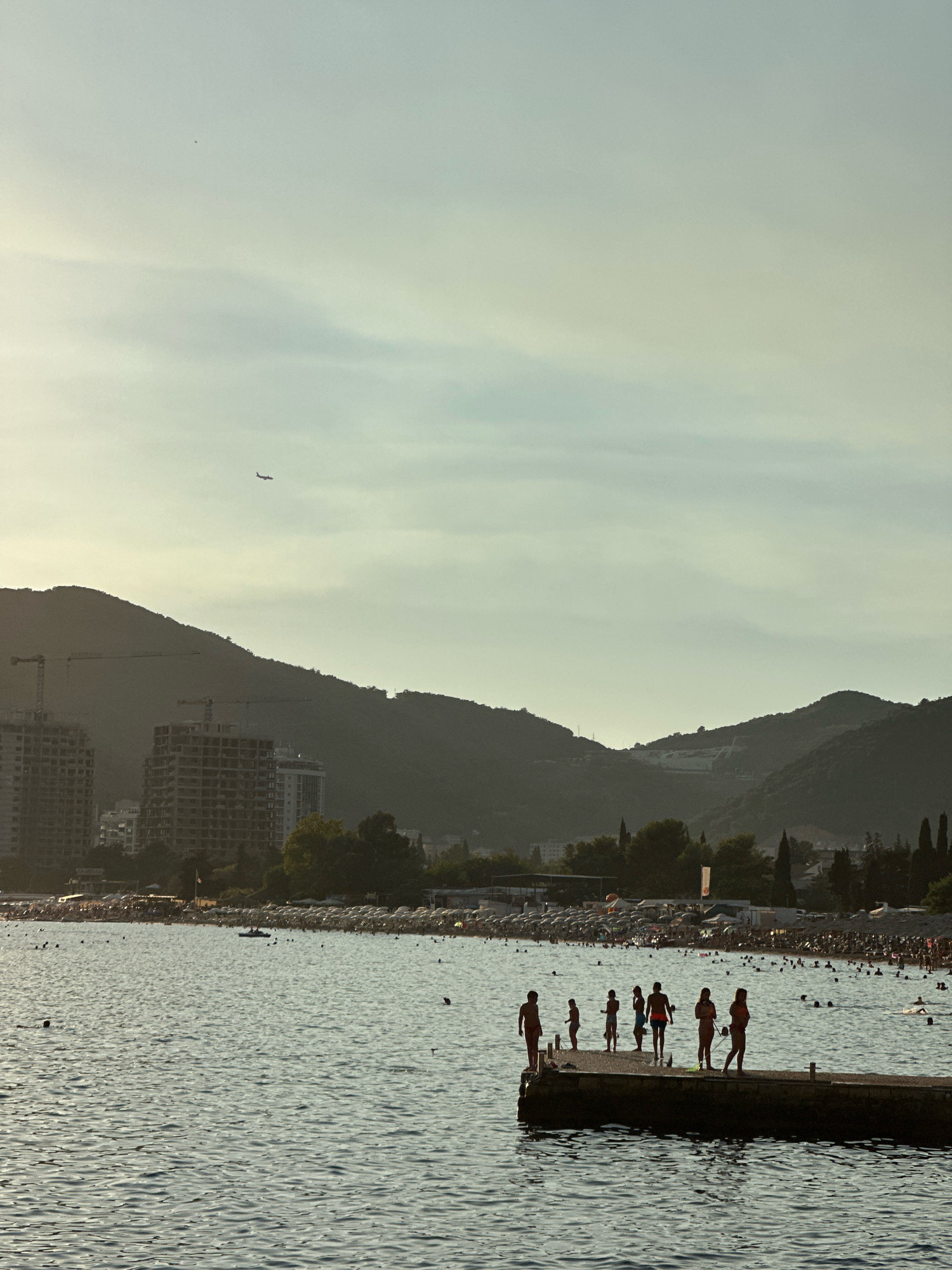 The image captures a serene coastal scene in Budva, Montenegro, during what appears to be late afternoon or early evening. The foreground is dominated by the calm, rippling surface of the Adriatic Sea, reflecting the muted colors of the sky. A concrete pier juts out into the water, serving as a gathering spot for a group of silhouetted figures. Among them, individuals are standing, seemingly enjoying the view or preparing to enter the water. A few are also visible swimming or wading in the sea.
In the mid-ground, a bustling beach is depicted, filled with numerous people relaxing under a multitude of white umbrellas. The shoreline is lined with trees, and behind them, a cityscape emerges with buildings under construction, characterized by exposed concrete structures and cranes. Further in the background, a series of dark, undulating mountains form a dramatic backdrop to the scene, their contours softened by the atmospheric haze and receding light. High in the pale sky, a small airplane is visible, a faint silhouette against the diffused sunlight. The overall impression is one of a popular tourist destination, blending natural beauty with ongoing development.