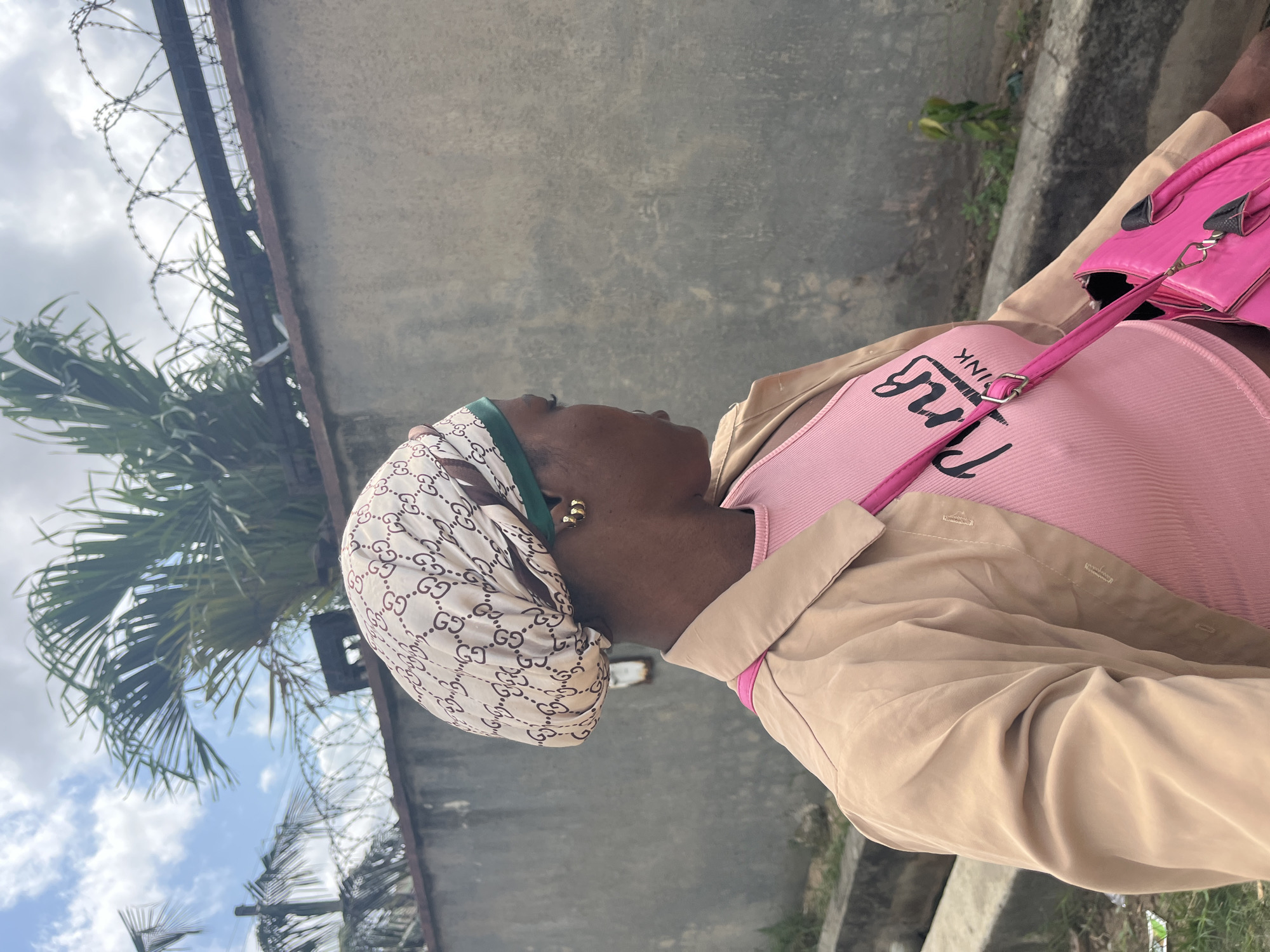 An African woman stands in the city of Ado, Nigeria. She wears a beige headscarf patterned with the Gucci logo, a pink top with the word "PINK" printed on it, and a tan jacket. She also carries a bright pink purse. Behind her is a tall, grey concrete wall topped with barbed wire, and a palm tree is visible. The sky is blue with scattered clouds, and there is vegetation growing alongside the walkway.