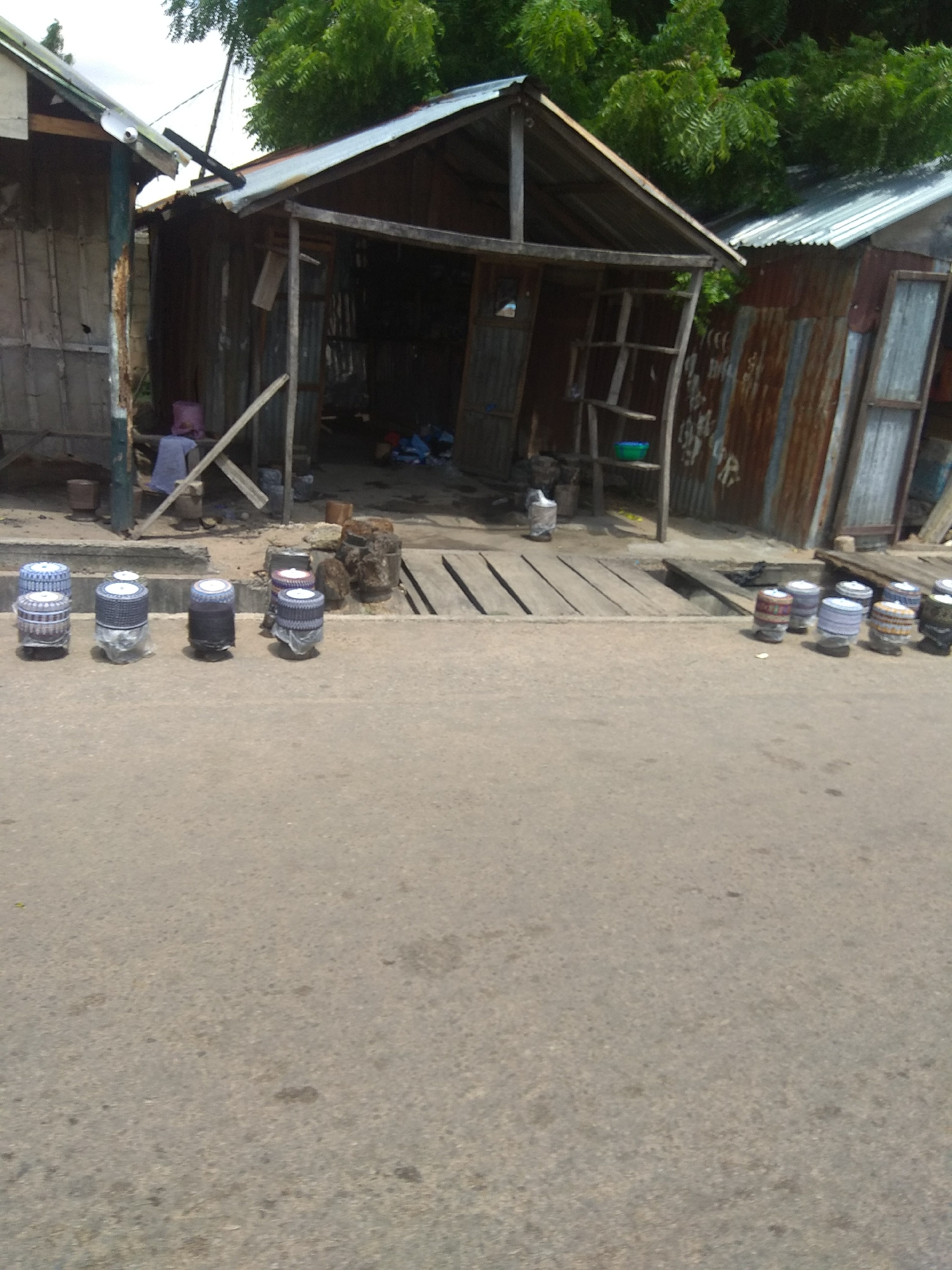 The image depicts a street scene in Jalingo, Nigeria. In the foreground, an asphalt road is visible, with a line of cylindrical objects, possibly hats or caps, displayed on the edge of the road. These objects are patterned with white and blue designs and some are partially covered in plastic. Behind the objects, a wooden walkway runs along the front of several informal structures. These structures appear to be market stalls or small shops, constructed from corrugated metal and wood, with open fronts. Some of these structures are dilapidated. In the background, lush green trees and foliage suggest a warm climate. The sky is overcast, indicating it may be daytime with diffused light. No people are clearly visible in the foreground or middle ground, though figures or activity might be obscured within the shaded interiors of the stalls. The overall impression is of a busy, informal marketplace. There is no visible text in the image.