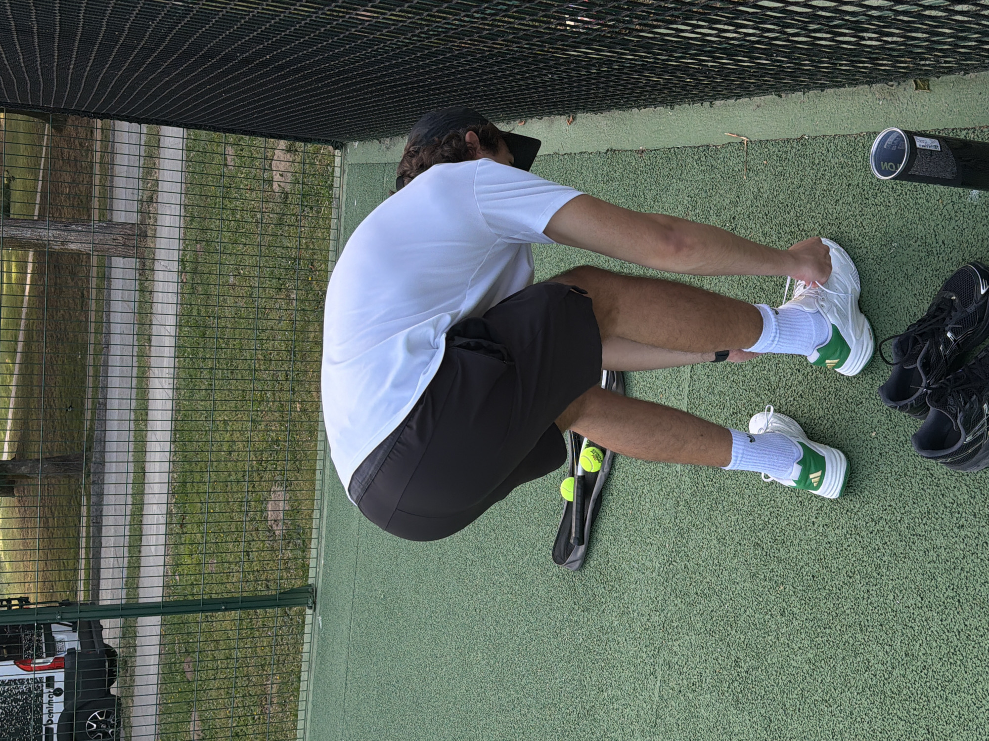 The image depicts a person wearing a white t-shirt, black shorts, and white and green athletic shoes on an outdoor green court. They are bending over near a black fence, with a tennis racket bag and two tennis balls on the ground beside them. In the background, a white van with text is visible behind a green metal fence, with trees and a pathway beyond that. The scene appears to be a sports facility, possibly a tennis court, in Santiago De Compostela, Spain.