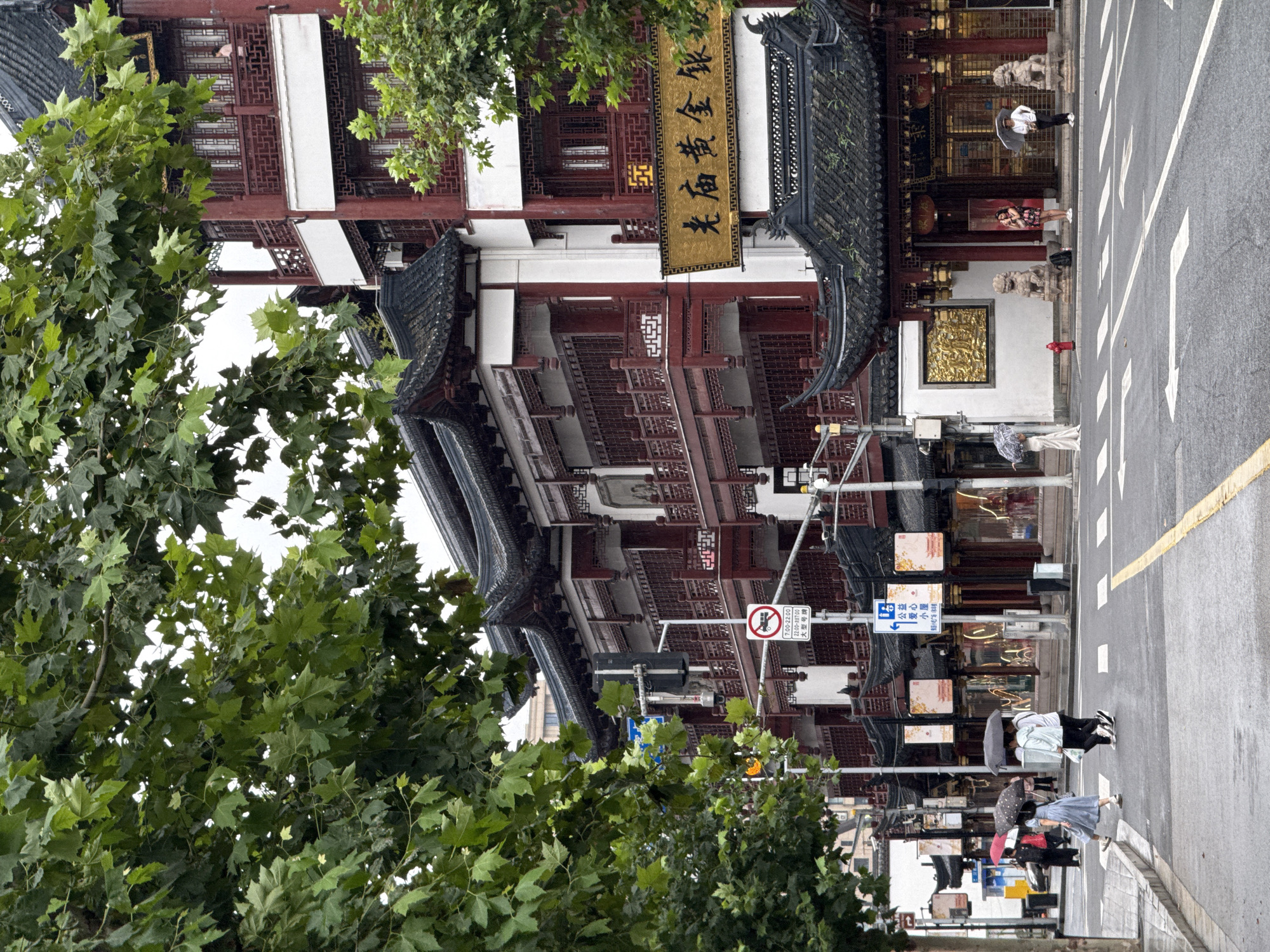 The image depicts a street scene in Shanghai, China, likely in the Yu Garden area, known for its traditional Chinese architecture. The foreground shows a wet asphalt street with white crosswalk markings and a yellow line dividing traffic lanes. A traffic light hangs overhead, and a "no buses" sign is visible on a pole.
On the right side of the street, a multi-story building features traditional Chinese architectural elements, including dark tiled roofs with upturned eaves, red wooden balconies with intricate latticework, and white walls. A large golden sign with Chinese calligraphy hangs on the building. Two stone lion statues guard the entrance to what appears to be a shop or restaurant.
Several people are present in the scene. Two individuals are walking across the street, one holding a white umbrella and the other a black umbrella. Another person, wearing a light blue dress, walks across the street holding a polka-dot umbrella. Further in the background, more people are visible on the sidewalk, some holding umbrellas.
On the left side of the street, a large tree with green leaves partially obscures the view of the buildings. The sky appears overcast, suggesting a cloudy or rainy day, which is consistent with the presence of umbrellas and the wet street. The overall atmosphere of the image is one of urban life set against a backdrop of historic architecture.