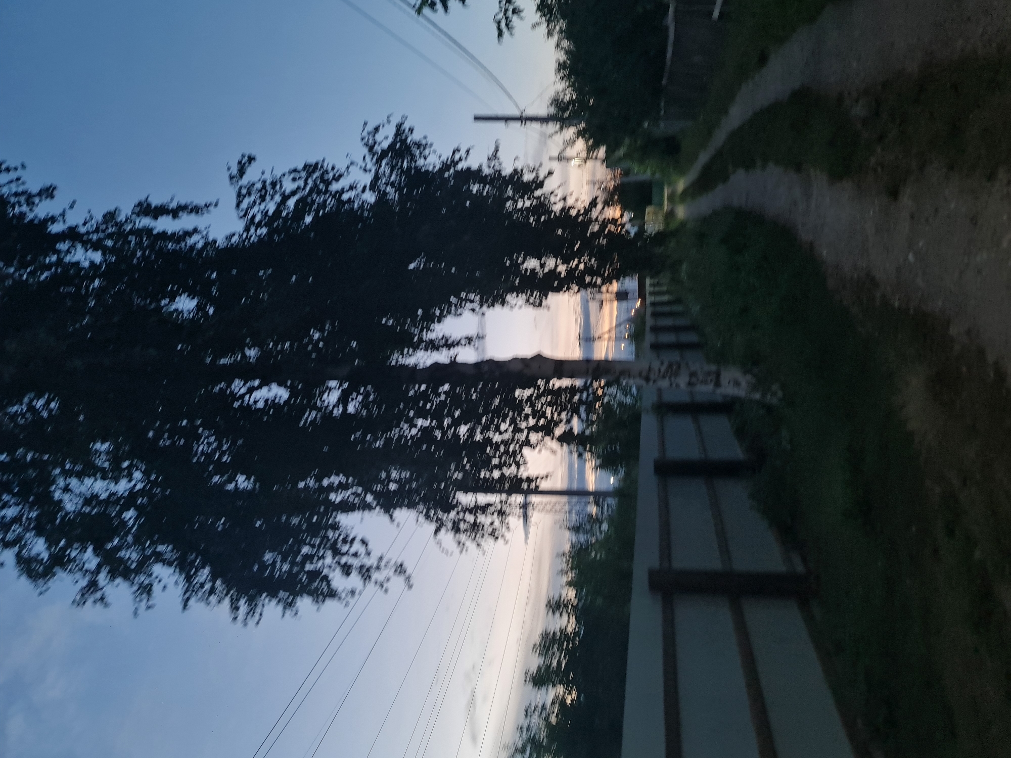 The image depicts a rural road at dusk in Angarsk, Russia, likely during late spring or summer. The sky is a gradient of deep blue and pale pink, indicating twilight. Several power lines crisscross the sky, supported by utility poles visible in the distance. On the left side of the road, a light-colored fence made of corrugated metal panels with dark wooden posts runs along the property line. Beyond the fence, there is dense green foliage and several trees, including a tall birch tree in the foreground with its dark silhouette against the sky. The road itself is unpaved, appearing to be a dirt or gravel path, with tire tracks suggesting it is used by vehicles. The overall scene is tranquil and natural, characteristic of a suburban or rural area. There are no people visible in the image, and the focus is on the landscape and the road.