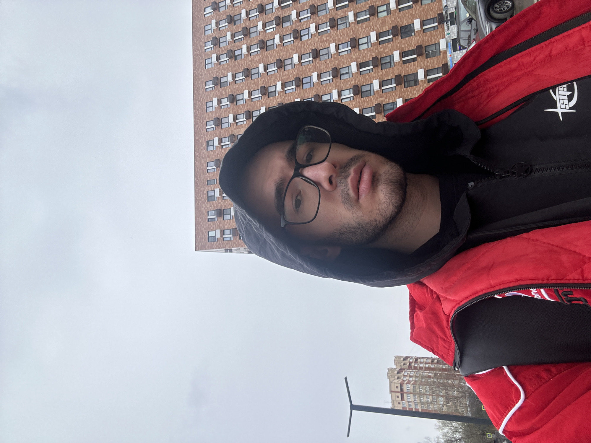 A man with glasses and a hooded jacket takes a selfie outside, with a tall residential building behind him. The location appears to be Moscow, Russia, based on the architectural style of the building, which is typical of Soviet-era construction. The man is wearing a black hooded sweatshirt under a red puffer vest. The sky is overcast, suggesting a cool or cold climate. In the background, there are other buildings and a street with parked cars. A lamppost is also visible in the bottom part of the image.
