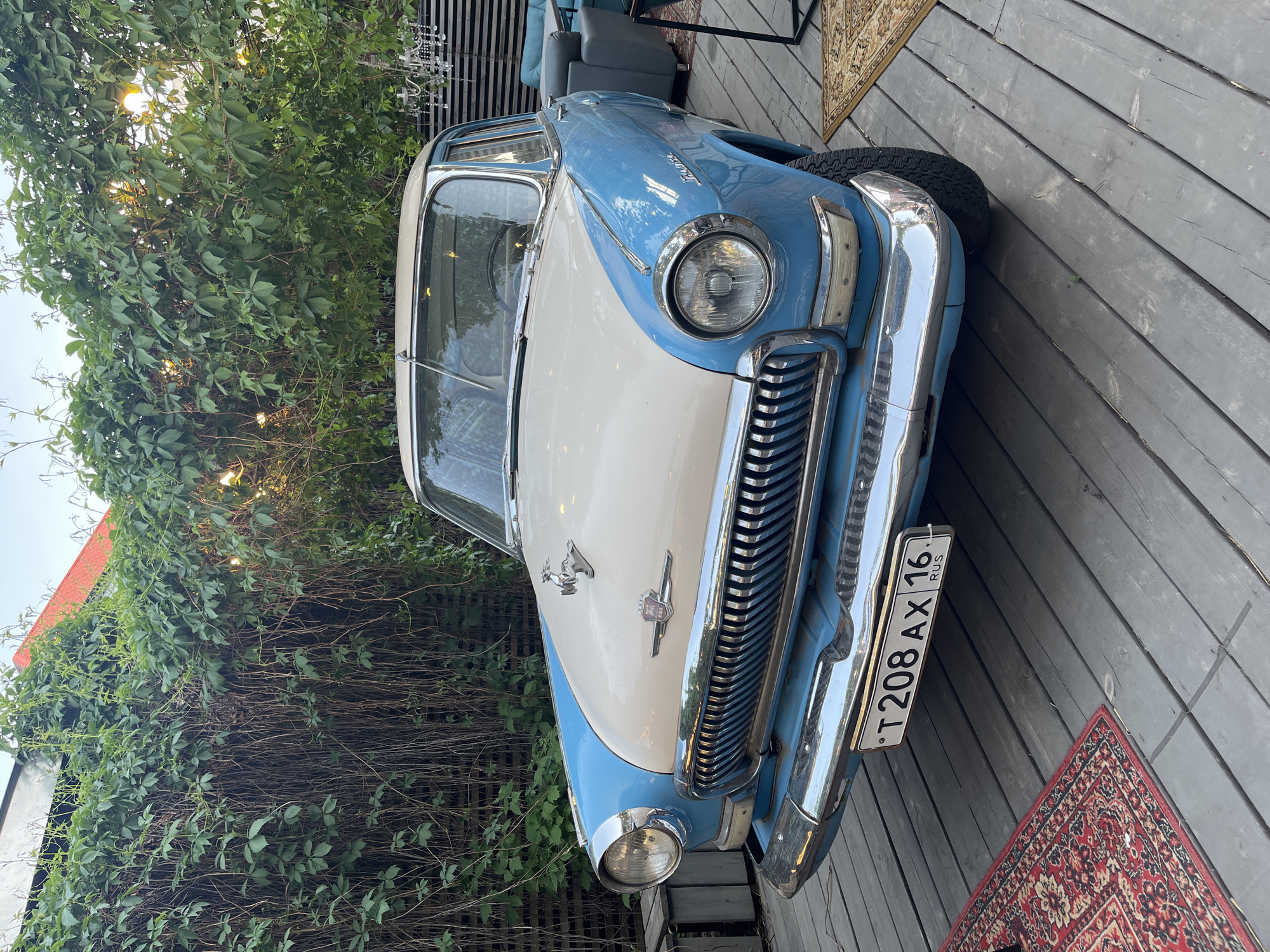 A two-tone blue and cream vintage car is parked on a wooden deck. The car has a license plate that reads "T208 AX 16 RUS". The car is located near a building covered in green vines. Two rugs are visible near the car. The setting suggests an outdoor cafe or restaurant. The license plate indicates the car is registered in Russia, possibly in the city of Kazan, where area code 16 is used. No people are visible in the image.