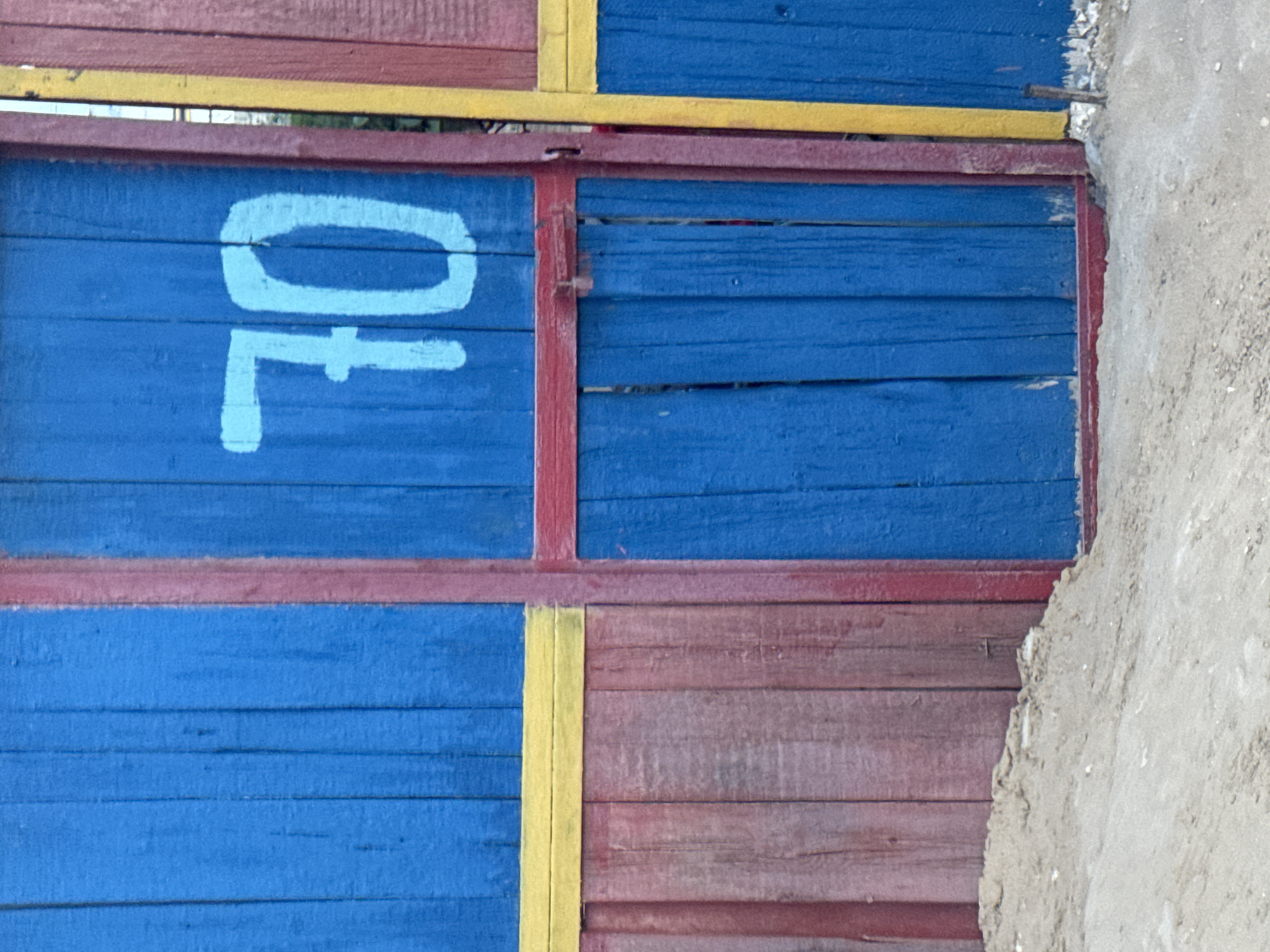 The image shows a close-up view of a brightly painted wooden fence, likely part of a private property or a recreational area in Atameken, Kazakhstan. The fence is divided into sections of varying colors: a prominent bright blue door with the number "70" stenciled in white is centrally located. To the left of the blue section, there is a segment painted in alternating blue and a duller, weathered reddish-brown wood. A horizontal yellow stripe runs across the lower portion of this blue and red section, separating it from the lower part of the fence which is entirely in the reddish-brown wood. To the right of the blue door, a vertical red beam borders it, followed by a narrow yellow stripe and then more reddish-brown painted wood. The fence is grounded in a patch of sand or gravel, suggesting an outdoor setting, possibly near a beach or a dusty area. The paint appears somewhat worn and chipped in places, indicating exposure to the elements. There are no people or animals visible in the frame. The scene is static and focuses solely on the architectural detail of the fence.
