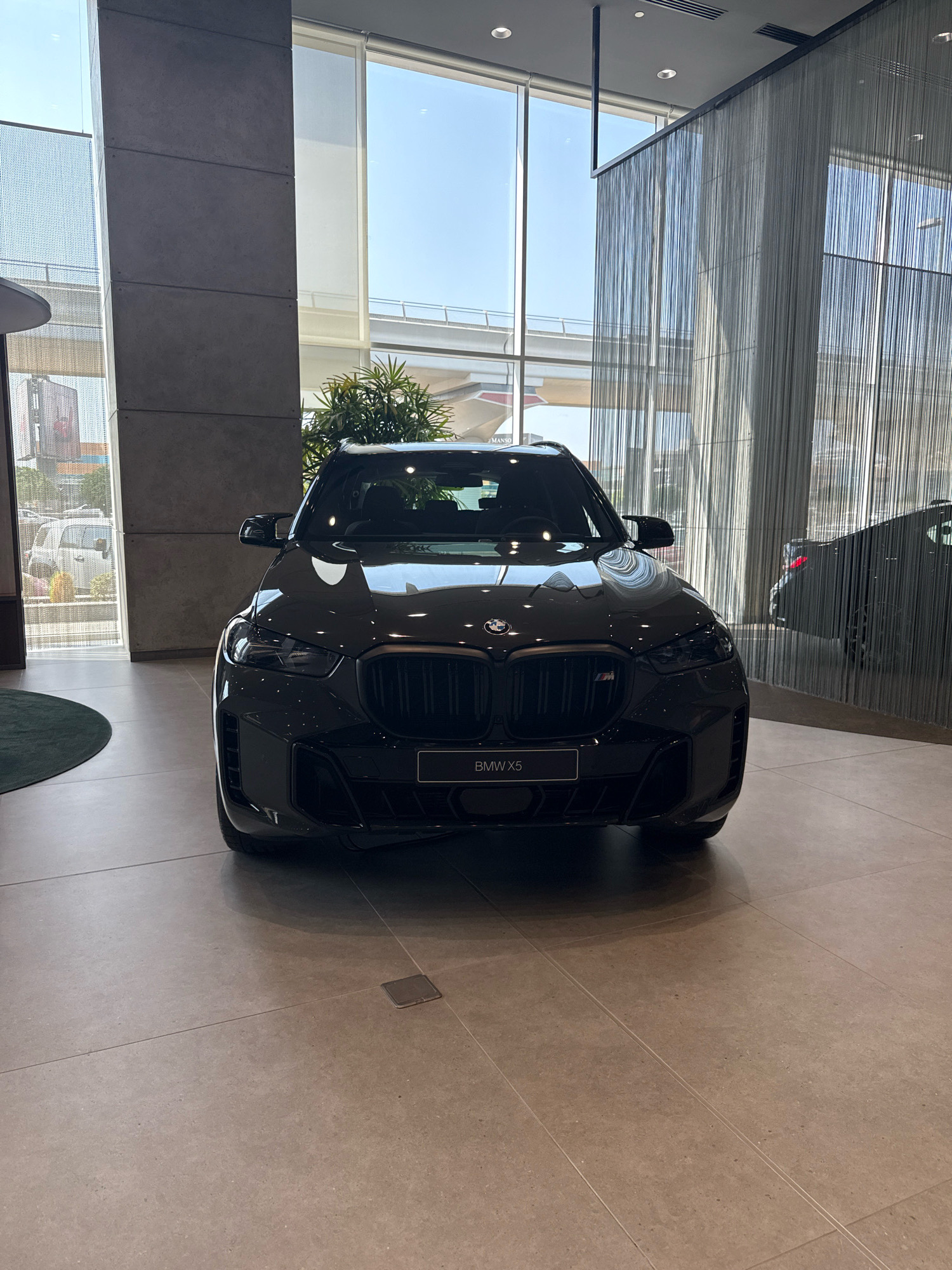 An eye-level, full frontal shot captures a dark-colored BMW X5 positioned centrally within a modern showroom. The vehicle, the primary subject, is sleek and imposing, with its distinctive grille and headlights prominently displayed. The showroom's interior features polished concrete flooring and large windows that allow ample natural light to flood the space, creating a bright and airy atmosphere. Behind the BMW X5, a second dark-colored vehicle, possibly another BMW, is partially visible, suggesting a car dealership. The background includes a sleek, modern architectural design with vertical metal strands that create a shimmering effect, reflecting the light and adding a touch of sophistication. The location is the BMW Showroom in Dubai, United Arab Emirates, as indicated by the signage on the car. The scene is clean and well-lit, highlighting the premium nature of the vehicles and the showroom's design.