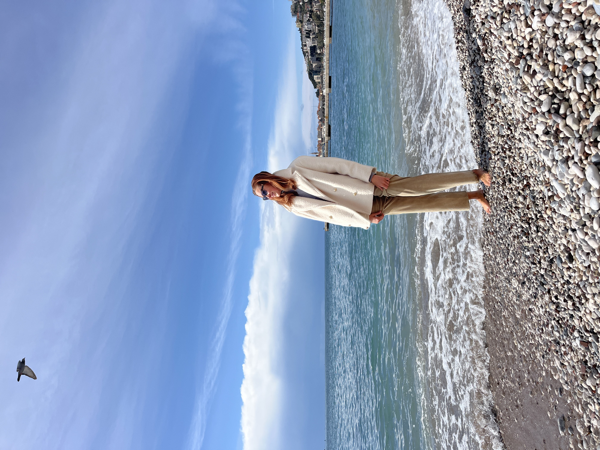 A daytime, eye-level photograph shows a single individual standing barefoot on a pebble beach in Budva, Montenegro, facing slightly left and looking towards the right. The person, with reddish-brown hair, wears sunglasses, a cream-colored double-breasted peacoat with gold buttons, and light brown corduroy trousers. Their bare feet are partially submerged at the water's edge, where small waves with white foam break on the multi-toned pebble shore. The turquoise-blue sea occupies the mid-ground, containing several dark, rectangular pier-like structures. In the background, the coastal town of Budva is visible, with numerous light-colored buildings and red roofs ascending a green hillside. The sky is predominantly blue with scattered white clouds, including a larger, denser cloud bank on the horizon above the sea. A dark-colored bird is in flight in the upper left portion of the sky.
