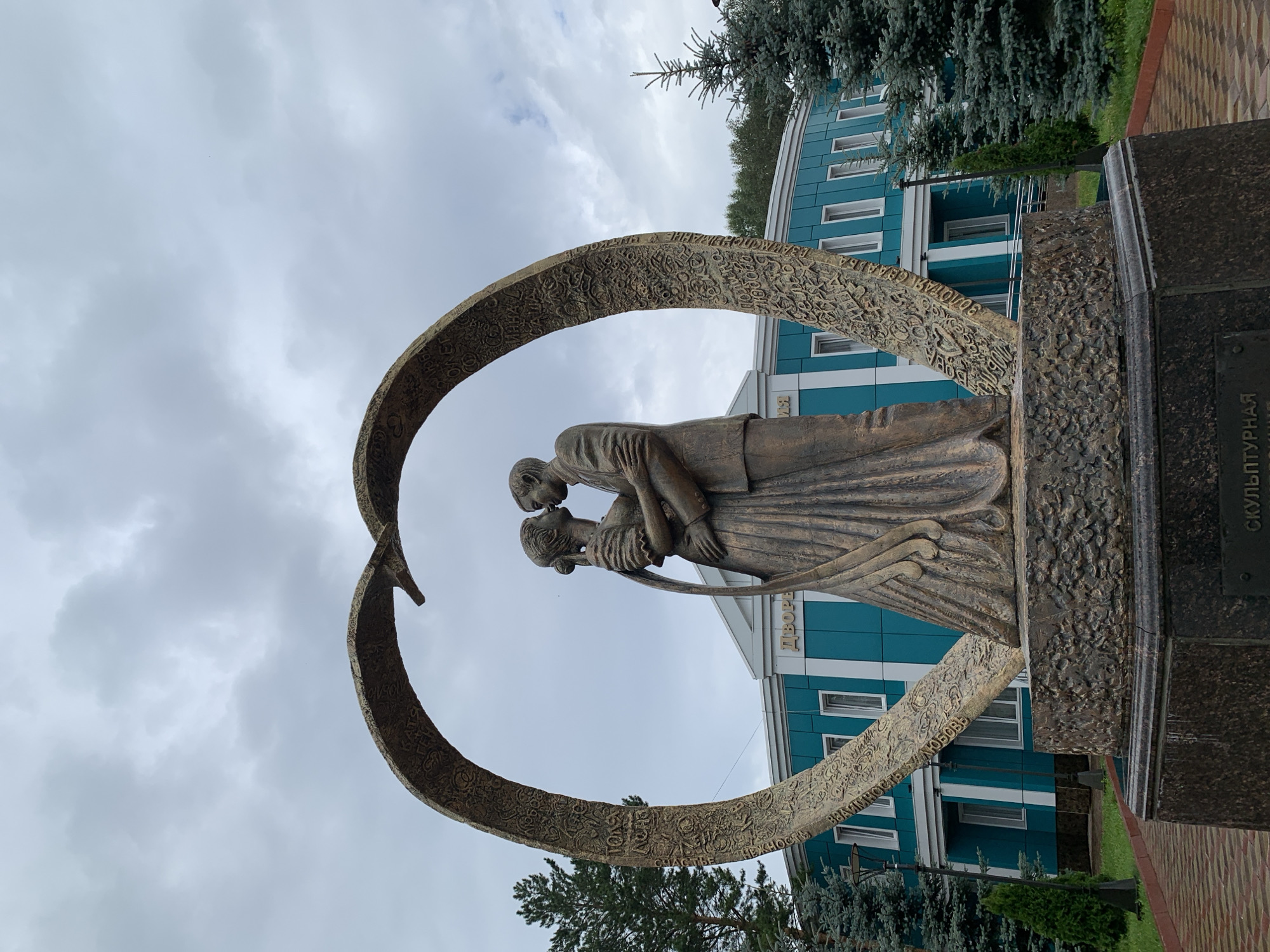 This is a full-shot, eye-level, outdoor photograph of a bronze sculpture of a couple embracing, framed by a large, ornate heart-shaped arch. The sculpture is positioned in front of a teal-colored building with white trim and windows. The building has Cyrillic text above its entrance. The sculpture depicts a man in a suit and a woman in a flowing wedding dress, kissing. The arch is textured with intricate carvings and text. Evergreen trees flank the sculpture on both sides. The sky is overcast. The sculpture is located in Prokopyevsk, Russia, in front of the Palace of Culture of the city.