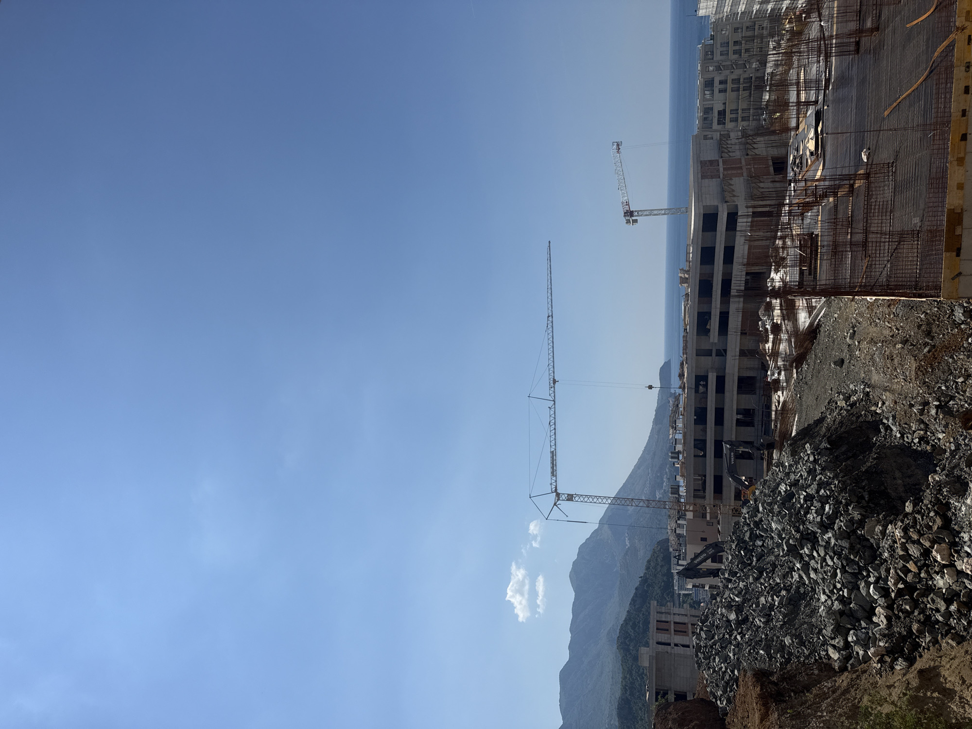 The image depicts a construction site in Boreti, Montenegro, under a clear blue sky. In the foreground, a large pile of grey rocks is visible on the left, with a construction site extending towards the right. Two large cranes dominate the mid-ground, with one tall tower crane on the left and a shorter one on the right, both actively participating in the construction of several multi-story buildings. These buildings appear to be in various stages of completion, with some showing concrete structures and exposed rebar, while others are nearing completion with exterior walls. In the background, rolling green hills and mountains rise, leading down to the shimmering blue sea. The presence of construction equipment like excavators, identified by the "VOLVO" brand on their arms, further emphasizes the ongoing construction activity. The scene suggests a developing area with new residential or commercial properties being built along the Montenegrin coast.