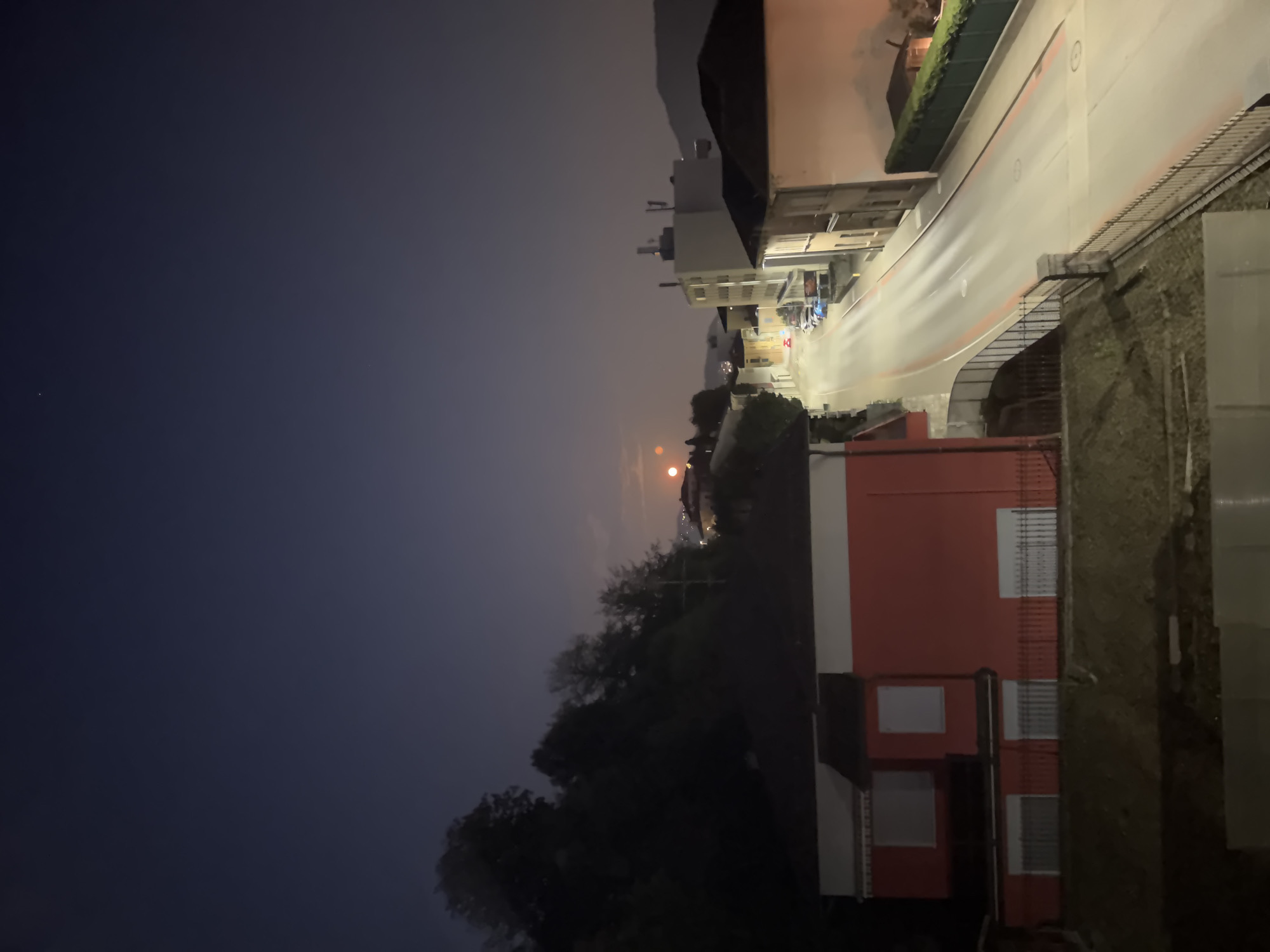 An elevated, nighttime view of a street in Balerna, Switzerland. The street is paved and lined with buildings on both sides. The sky is a dark blue, with a faint glow on the horizon and a bright object, likely the moon, visible in the upper left quadrant of the scene. Several cars are parked along the street, and a few are in motion, indicated by light trails. There are trees on a hillside to the left of the street, and a red building with white-trimmed windows is prominent in the foreground on the left. The overall scene depicts a quiet, urban street at night, illuminated by artificial light and the moon.
