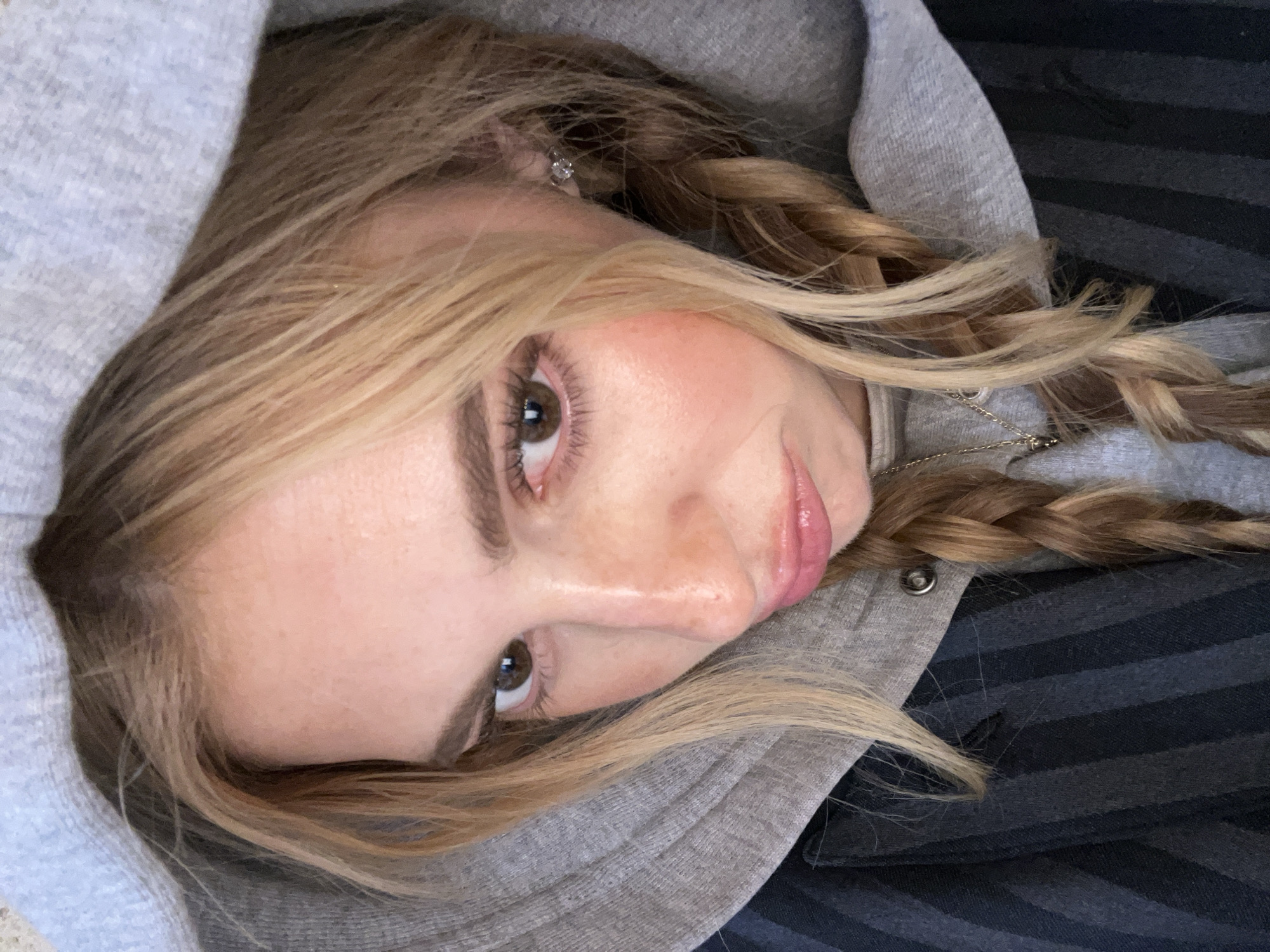 A close-up portrait shows a young woman with fair skin and light brown hair styled in two braids. She is wearing a grey hooded sweatshirt with the hood partially obscuring her forehead. Her eyes are light brown, and she has a slight smile. She has a small earring in her left ear. Part of the background reveals a dark, striped fabric. The image provides no information to indicate it was taken in Paris, France.