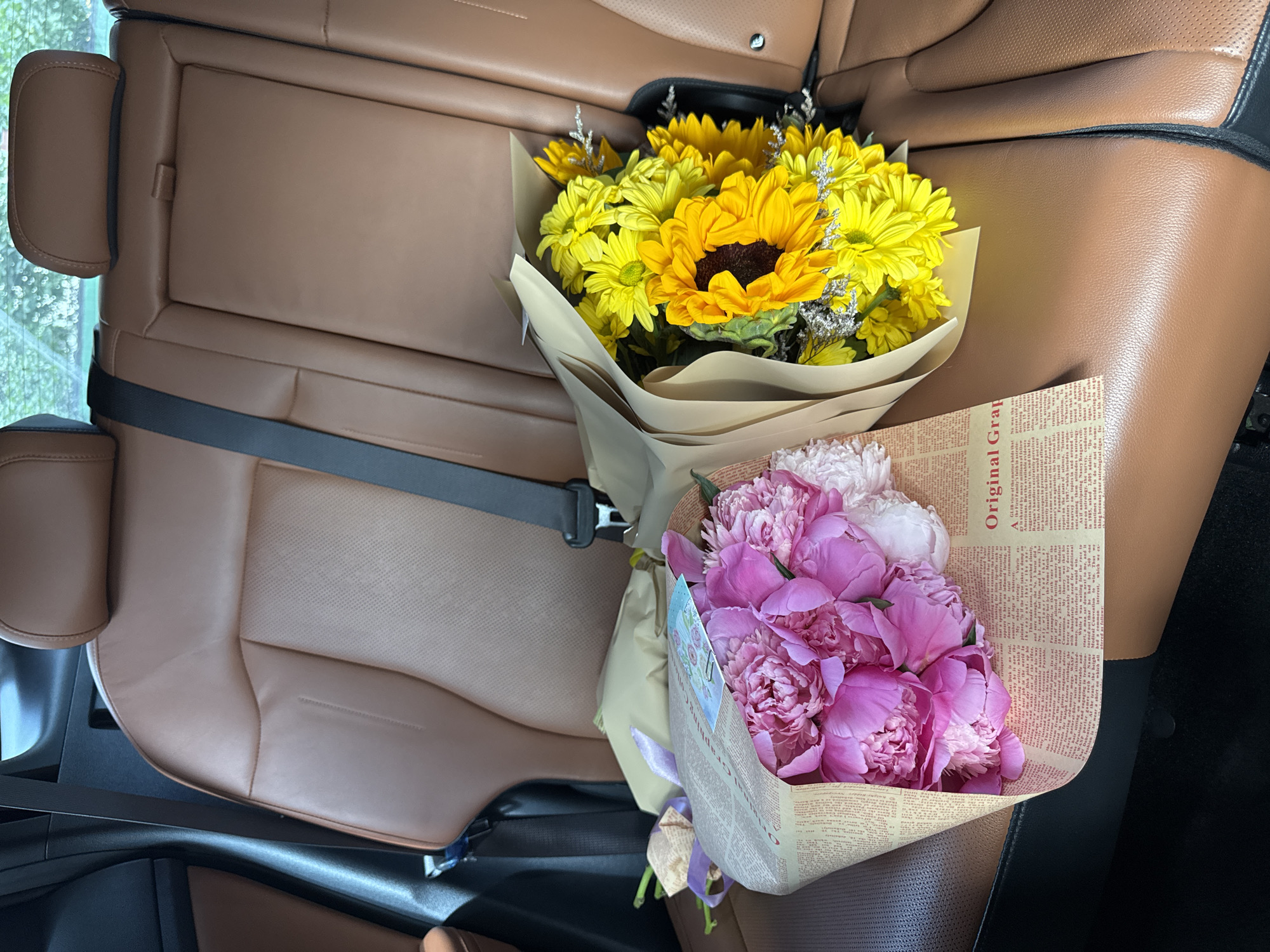 Two bouquets of flowers, one with pink peonies and the other with sunflowers and yellow daisies, are placed on the leather seats of a car, likely in Almaty, Kazakhstan. The car's interior is visible, featuring brown leather seats and seatbelts. The bouquets are wrapped in paper, with the pink bouquet also having a purple ribbon. The scene appears to be captured from a medium shot, focusing on the flowers and the car's interior. The background is blurred, suggesting an outdoor environment, possibly with trees visible through the car's windows.