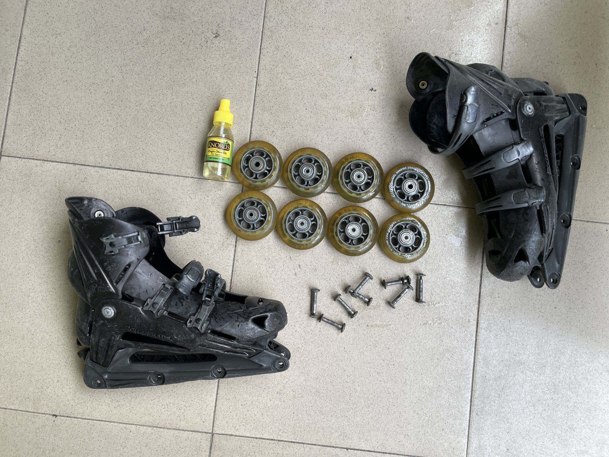 The image shows a collection of rollerblading equipment laid out on a tiled floor. In the upper left quadrant, there is a black rollerblade boot and frame. To the right of this, there is a small yellow bottle of oil with a white label that reads "KNORRIS" and "Tiger Stripe Oil". Below the oil bottle, there are eight rollerblade wheels arranged in two columns of four. Scattered among the wheels and to the left of them are several silver screws. In the bottom right quadrant, there is another black rollerblade boot and frame. The scene is consistent with a disassembled rollerblade, likely being maintained or prepared for use. The tiled floor suggests an indoor environment, possibly a garage or storage area, within the city of Ado, Nigeria. There are no people or animals present in the image.