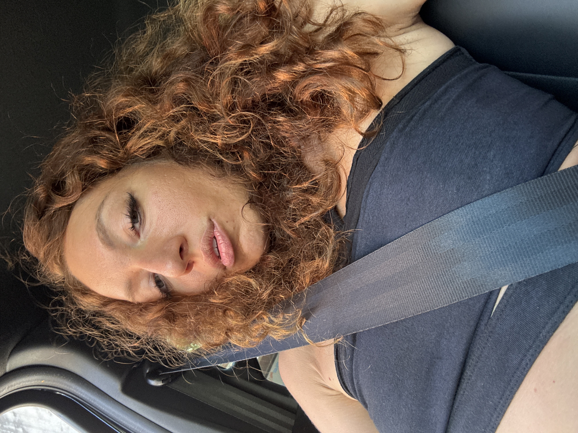 A woman with curly, reddish-brown hair is sitting in a vehicle, wearing a dark blue tank top and a gray seatbelt. She is looking down and to the side with a somewhat wistful expression. The interior of the car is dark, and the window is visible in the lower left corner of the image. The scene appears to be in an area with overcast lighting, as evidenced by the window view. There is no specific indication that this photo was taken in Sa Pa Ward, Vietnam.