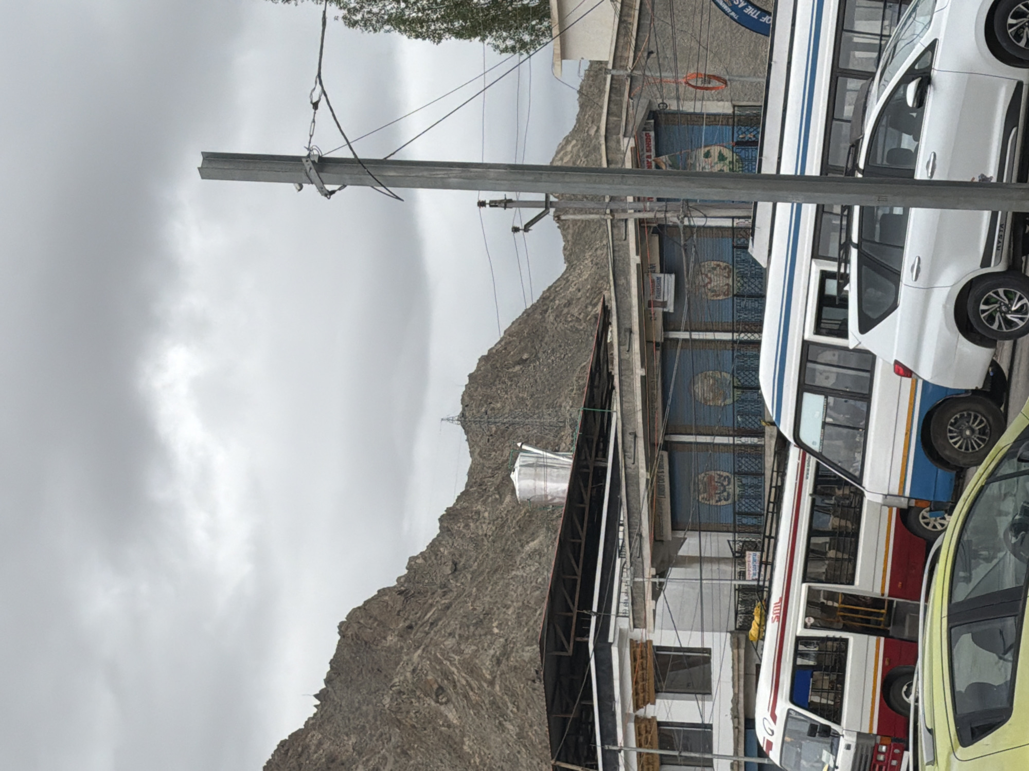 The image depicts a street scene in Leh, India, with a backdrop of rugged, brown mountains under a cloudy sky. In the foreground, several vehicles are parked and in motion. Two buses are prominent: one is white with red and blue stripes, and the other is white with blue and yellow stripes. A yellow car is partially visible in the lower left corner, and a white SUV is positioned behind the buses.
Buildings line the street, featuring traditional architecture with white facades, wooden window frames, and dark roofs. One building has a large silver water tank on its roof. Roller shutters on some of the shopfronts are painted with circular designs. Electrical poles with numerous wires crisscross the scene, indicating urban infrastructure. The overall atmosphere is that of a bustling town on a somewhat overcast day, with the natural landscape of the Himalayas dominating the background.