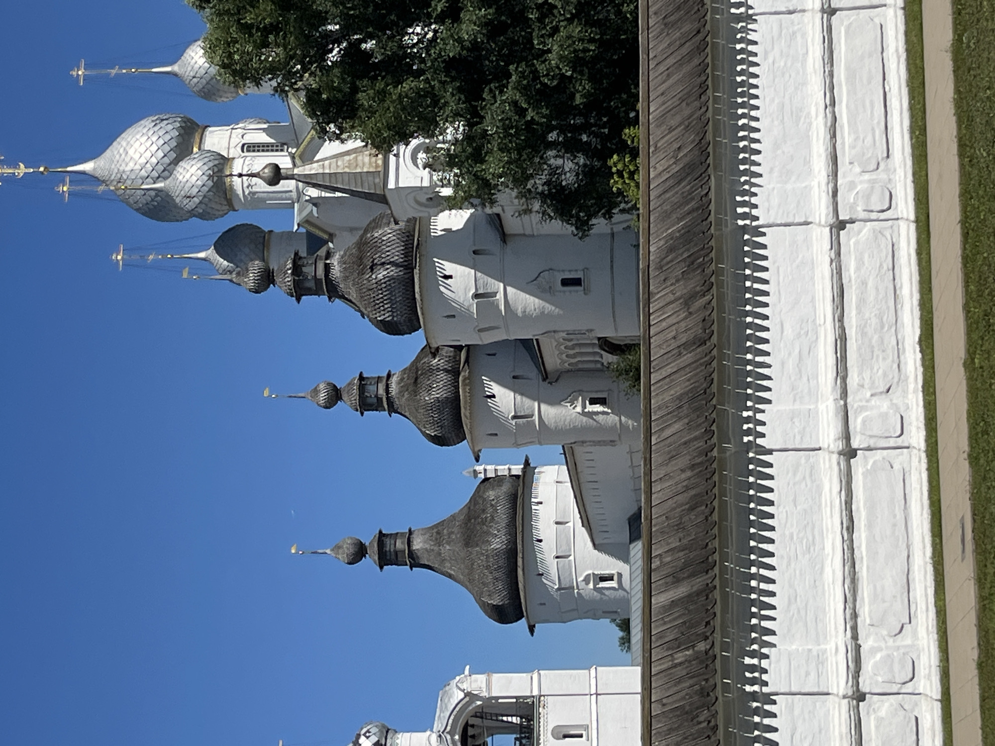 The image shows a view of the Rostov Kremlin in Rostov, Russia, on a clear, sunny day. The foreground features a white, textured wall with a wooden roof, and a neatly trimmed green lawn and paved walkway. Behind the wall, several white buildings with dark, onion-shaped domes rise against a bright blue sky. One prominent structure on the left is a bell tower with a white archway. The architectural style suggests a Russian Orthodox cathedral complex. The lighting indicates it is daytime, likely late morning or early afternoon due to the shadows. No people are visible in the frame.