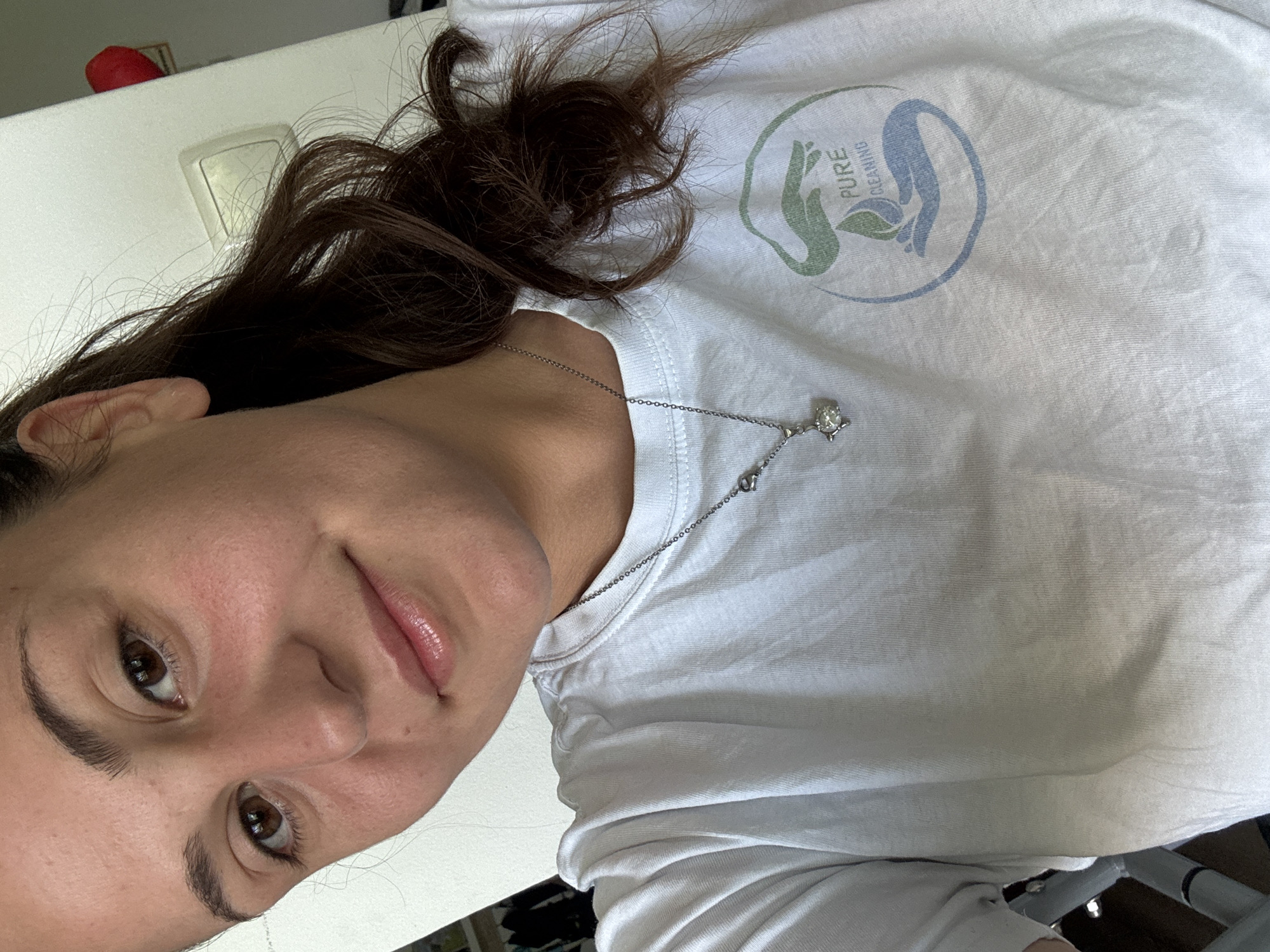 The image shows a close-up, selfie-style shot of a young woman with brown hair, brown eyes, and fair skin. She is wearing a white t-shirt with a circular logo on the right side of her chest. The logo features two hands, one in green and one in blue, with a green leaf between them, and text that reads "PURE CLEANING". A silver necklace with a small, sparkly pendant is visible around her neck. She is smiling slightly, and her hair is pulled back. The background is out of focus, but appears to be an indoor setting with a light-colored wall and a light switch. There is a hint of a red object on the wall to the right. The image does not contain any discernible information that would place it in the city of Plovdiv, Bulgaria.