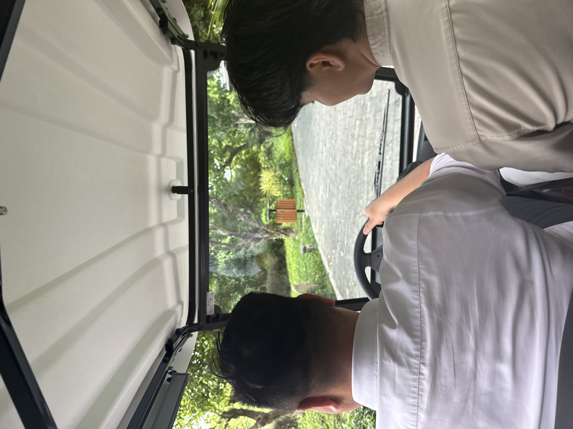 Two people are sitting in a golf cart. The golf cart is driving on a paved road with trees and grass on either side. In the background, there is a wooden bench. The image was likely taken in Nha Trang Ward, Vietnam. One person is driving the golf cart, and the other person is riding in the passenger seat. The driver is wearing a white shirt and the passenger is wearing a tan shirt. The golf cart has a white roof and black frame. A windshield wiper is visible in front of the driver.