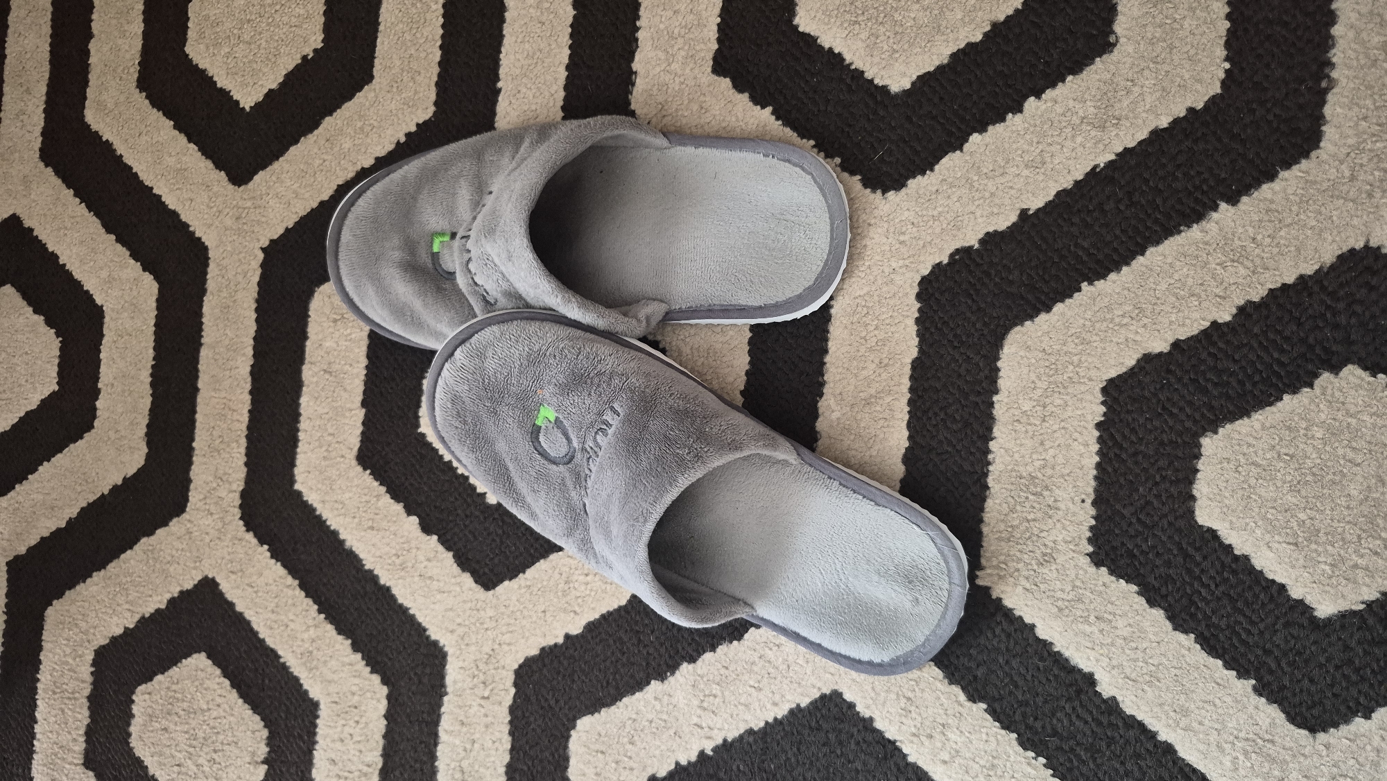 Two grey plush slippers are positioned on a rug with a black and beige geometric pattern. The slipper on the left is slightly angled, with its toe pointing towards the upper left of the frame. It has a logo embroidered on it, which appears to be a green symbol with some lettering underneath. The slipper on the right is positioned behind and to the right of the first, with its toe pointing towards the bottom of the frame. It also has a small green element visible on its upper surface, near the toe. The rug's pattern consists of thick, alternating bands of black and beige, forming a series of connected hexagonal shapes. The slippers are the primary subjects, and their soft texture and color are clearly depicted against the contrasting rug. The image appears to be taken from a top-down perspective. There is no indication of the slippers being in use or any human presence. The scene is static and presents a still life arrangement of household items. The provided information about the location in Krauja, Latvia, cannot be visually confirmed from the image itself.