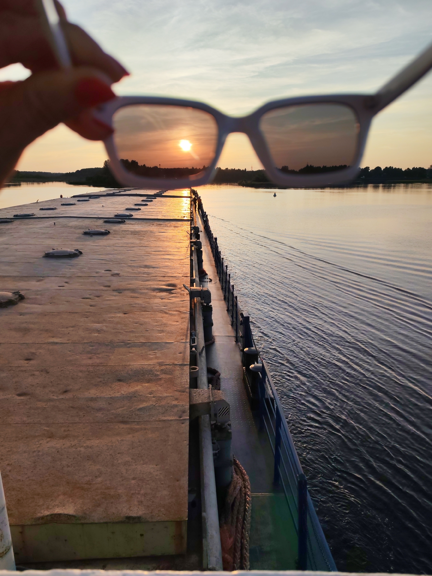 A person's hand, with red nail polish, holds up a pair of white-framed sunglasses, obscuring the view of the sunset over a body of water in Vytegra, Russia. The sunglasses reflect the setting sun, casting a warm orange glow. In the foreground, a portion of a boat's deck is visible, with a textured surface and several circular hatches. To the right, the boat's railing runs along the edge, with the dark water of the river creating ripples. The background shows a tree-lined horizon under a pale sky.