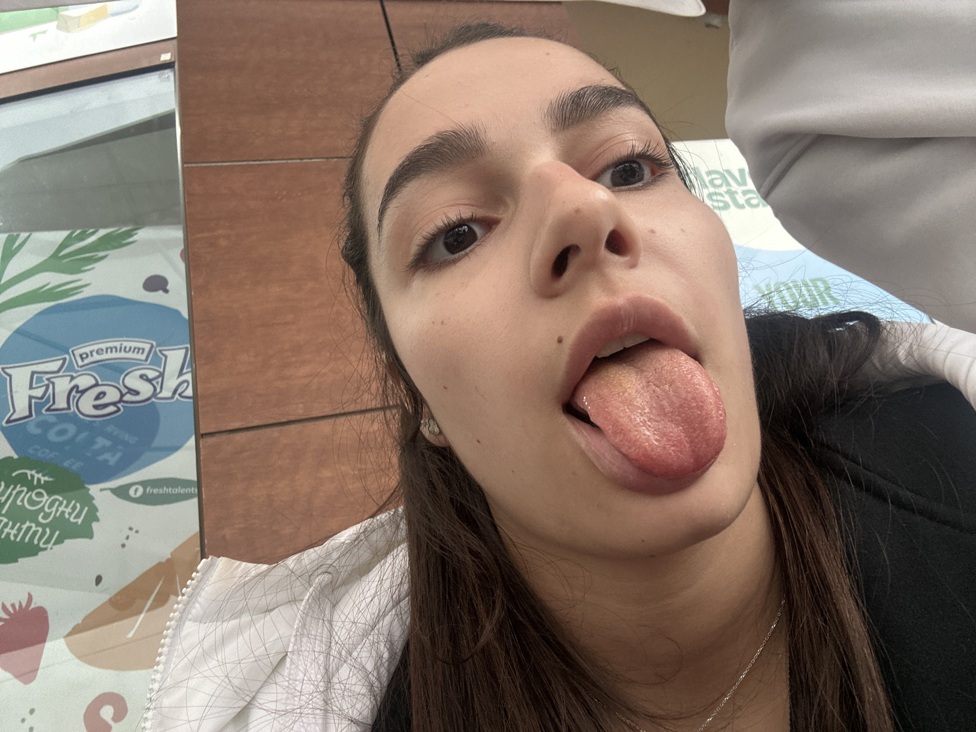 This close-up indoor shot features a young woman with dark hair and prominent eyebrows, captured from a low angle. She is sticking out her tongue, looking directly upwards towards the camera, conveying a playful or silly expression. Her dark hair is pulled back, and she is wearing a white zippered jacket over a black top, with a delicate silver necklace visible.
The background suggests a commercial setting, possibly a café or food establishment in Plovdiv, Bulgaria. To the left of the woman, a display or poster features the words "premium Fresh" and "COFFEE," along with the social media handle "@freshtalents." Partially visible Cyrillic text on the poster appears to read "ПРИРОДНИ" (Natural) and "НАПИТКИ" (Drinks), reinforcing the setting as a place selling beverages. A brown tiled wall is also visible in the background, to the right of the woman. The overall impression is an informal, humorous moment captured in a public space.