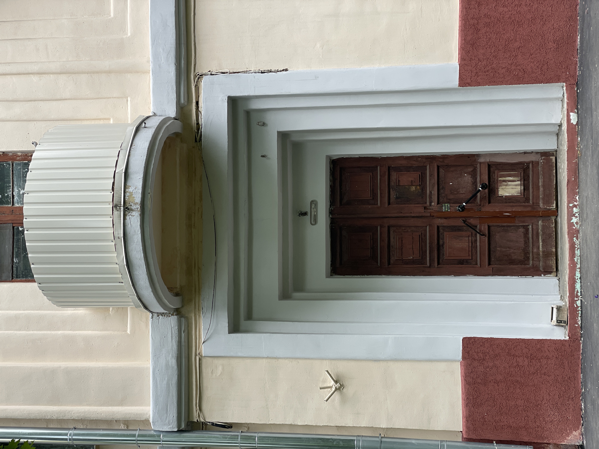 An eye-level, slightly upward shot reveals a doorway set within a layered, rectangular frame on the exterior of a building. The doorway is central, featuring a pair of aged, dark brown wooden doors, each composed of square panels, and a long, slender locking mechanism with black accents. The frame around the doorway is white, comprised of multiple receding layers, creating a tunnel-like effect.
To the left of the doorway, a corrugated metal structure curves outward, attached to the building. It is white with a gray border. Adjacent to this structure, a window with a brown frame is visible. The building's facade is primarily pale yellow, with areas of red brick visible on the right side of the doorway. Near the bottom of the image, a metallic pipe runs horizontally.
The scene is of an urban environment with some architectural elements. The location is the city of Khimki, Russia. No people are visible.