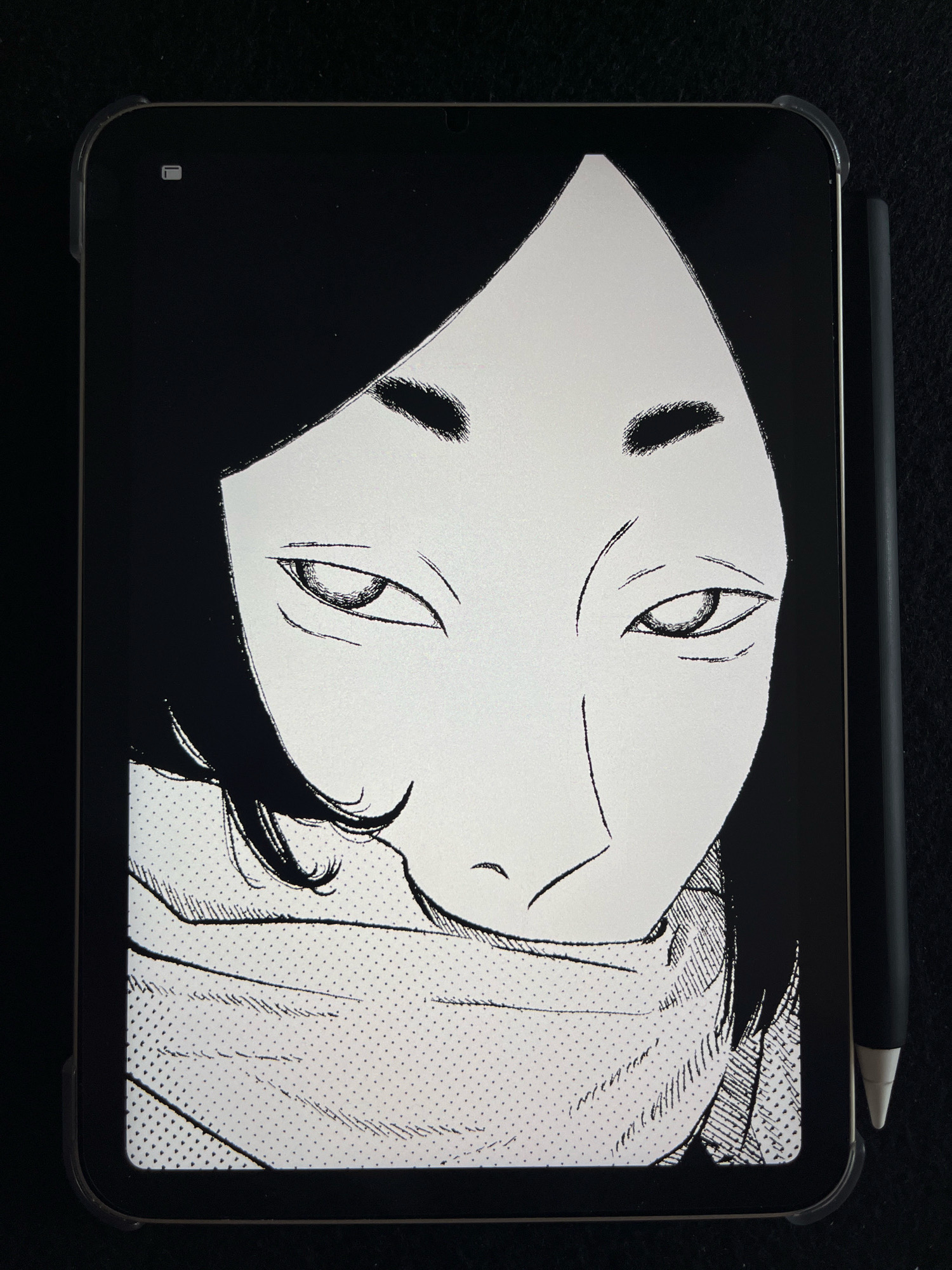 A close-up shot shows a tablet displaying a black and white illustration of a person's face. The artwork is in a manga style, with heavy lines and shading, depicting dark hair framing the face and a scarf or clothing around the neck. The person's eyes are looking slightly upwards, and their expression is neutral. The tablet is held in a clear case, and a stylus is positioned vertically to the right of the tablet, resting against its edge. The background is a dark, textured surface, likely fabric. There are no identifiable elements that would place this image in Wałcz, Poland.