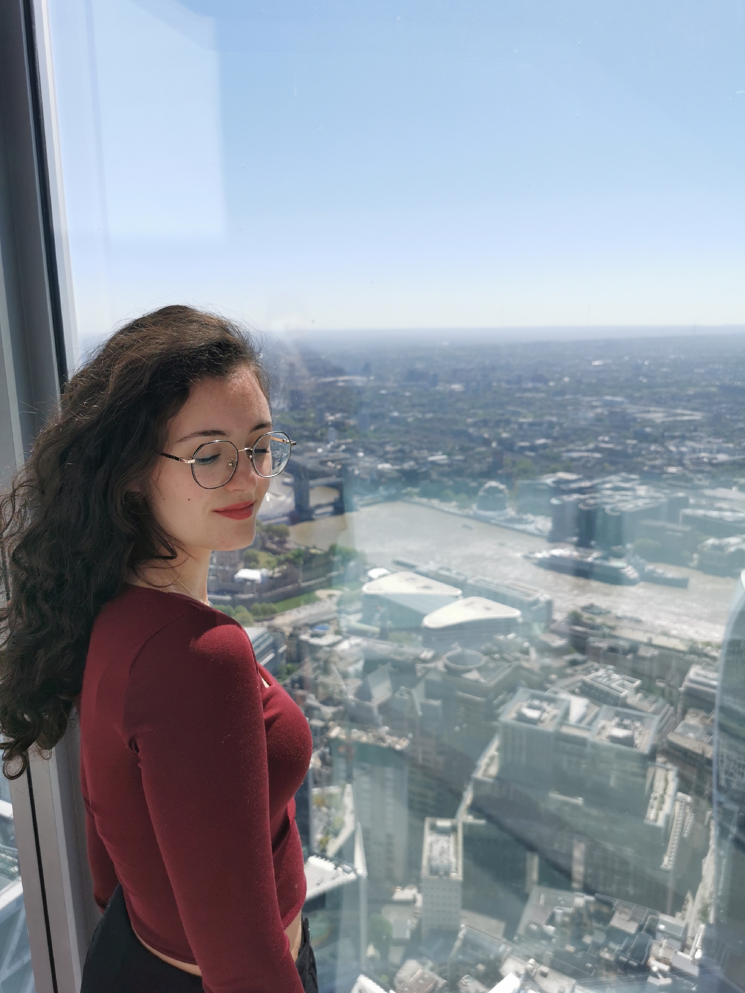 A young woman with long, dark, curly hair and round-framed glasses stands next to a large window, looking out over a sprawling cityscape. She is wearing a long-sleeved, deep red top and has red lipstick on. Her eyes are partially closed or looking down, and she has a gentle, contemplative expression on her face.
The setting is an interior space, likely an observation deck or high-rise building, overlooking a vast urban landscape. The bright outdoor light indicates it is daytime, probably a clear late morning or afternoon, though a slight atmospheric haze softens the distant view.
Beyond the window, a wide river, characteristic of the River Thames, winds through the heart of the city. Numerous buildings, ranging from modern structures to older architecture, stretch into the distance under a pale blue sky. The elevated vantage point offers an expansive view of London, United Kingdom, with its dense urban fabric visible for miles. There are no identifiable texts visible within the image. The overall mood conveyed is one of peaceful observation and appreciation of the impressive city panorama.