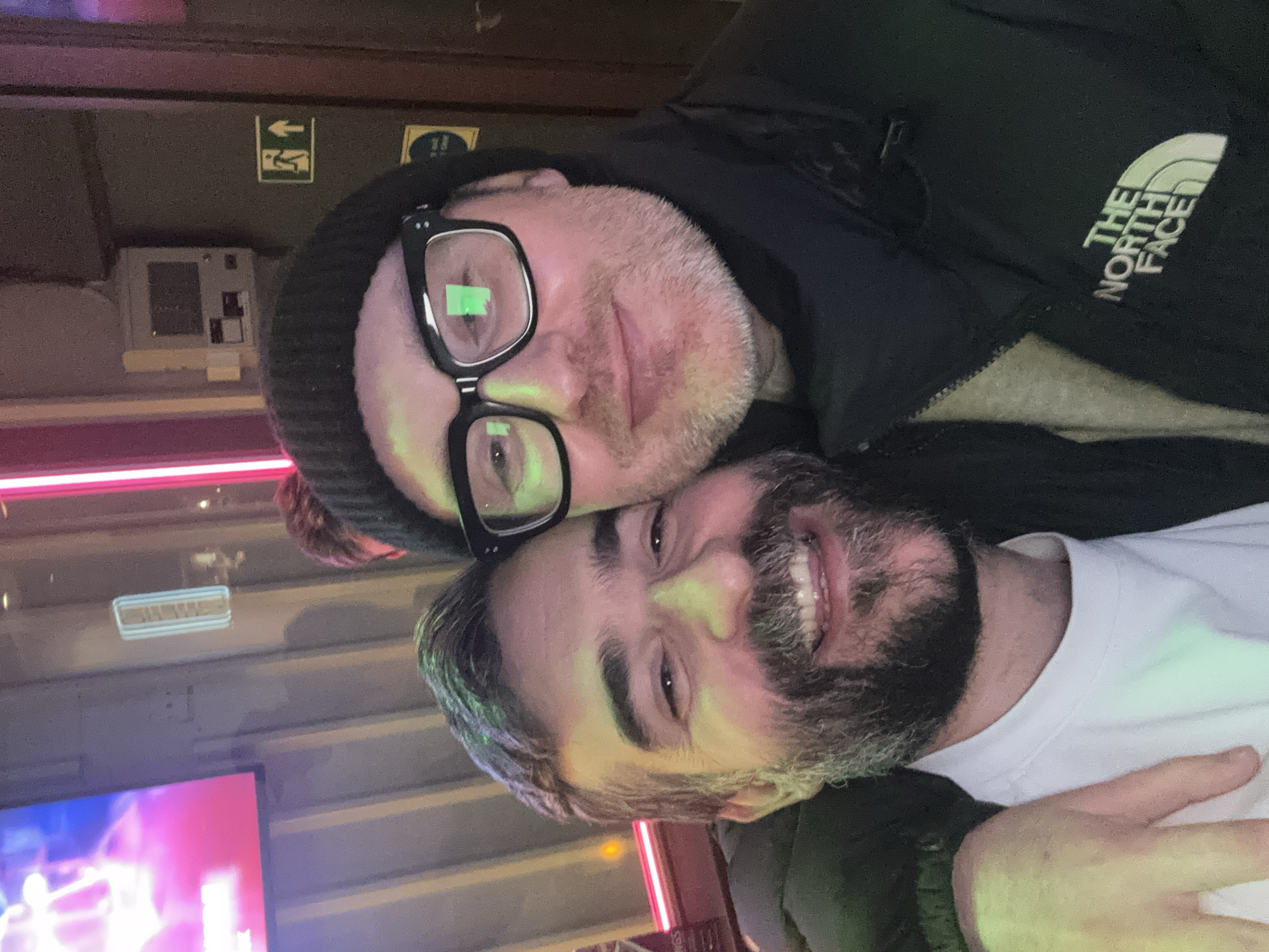 Two men are posing closely together for a photograph. The man in the foreground has dark hair, a full beard, and is wearing a white t-shirt. He is smiling and facing the camera. The other man behind him is wearing glasses and a black beanie hat. He has a lighter beard and is wearing a black jacket with "The North Face" logo on the sleeve. The setting appears to be indoors, likely a bar or pub, as suggested by the presence of a television screen in the background and interior lighting. The image was taken at The Phoenix pub on Cavendish Square in London.