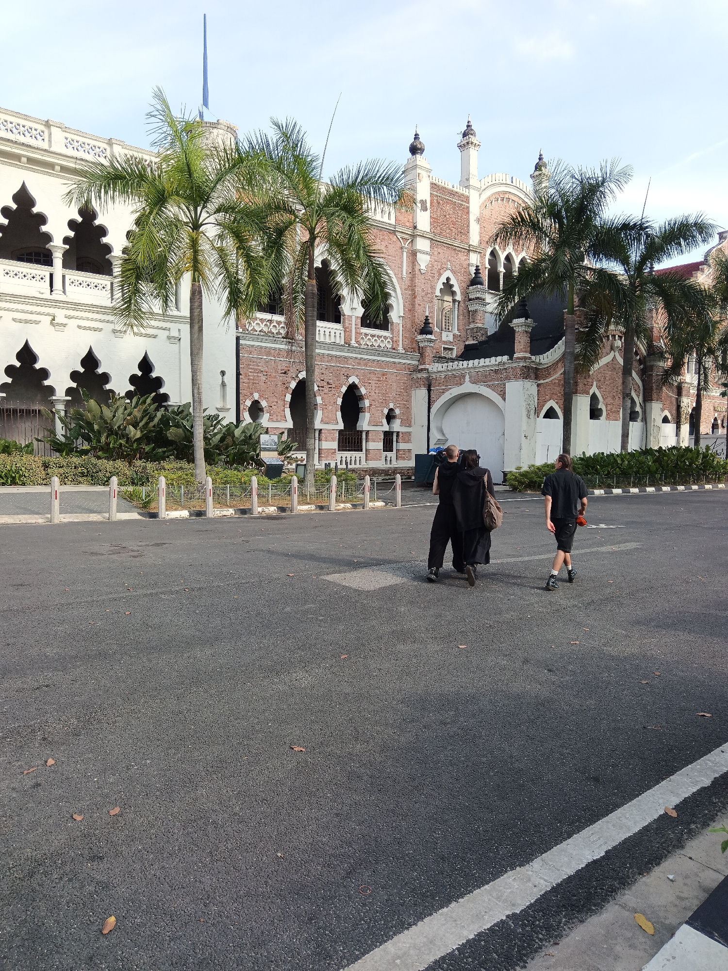 In Kuala Lumpur, Malaysia, in front of the Sultan Abdul Samad Building, three individuals walk across an asphalt road. The building, an iconic landmark constructed in the late 19th century, features distinctive Mughal-inspired architecture with a white facade and red brick sections. Two individuals, dressed in dark clothing and appearing to walk in unison, are in the foreground. The person on the left has their arm around the person in the middle, who is carrying a brown bag over their shoulder. A third individual, dressed in a black t-shirt and shorts, walks ahead of them. The scene is illuminated by daylight, with palm trees and lush greenery framing the foreground and the historic building in the background. The sky above is clear and light blue. The road shows signs of wear with some scattered leaves.