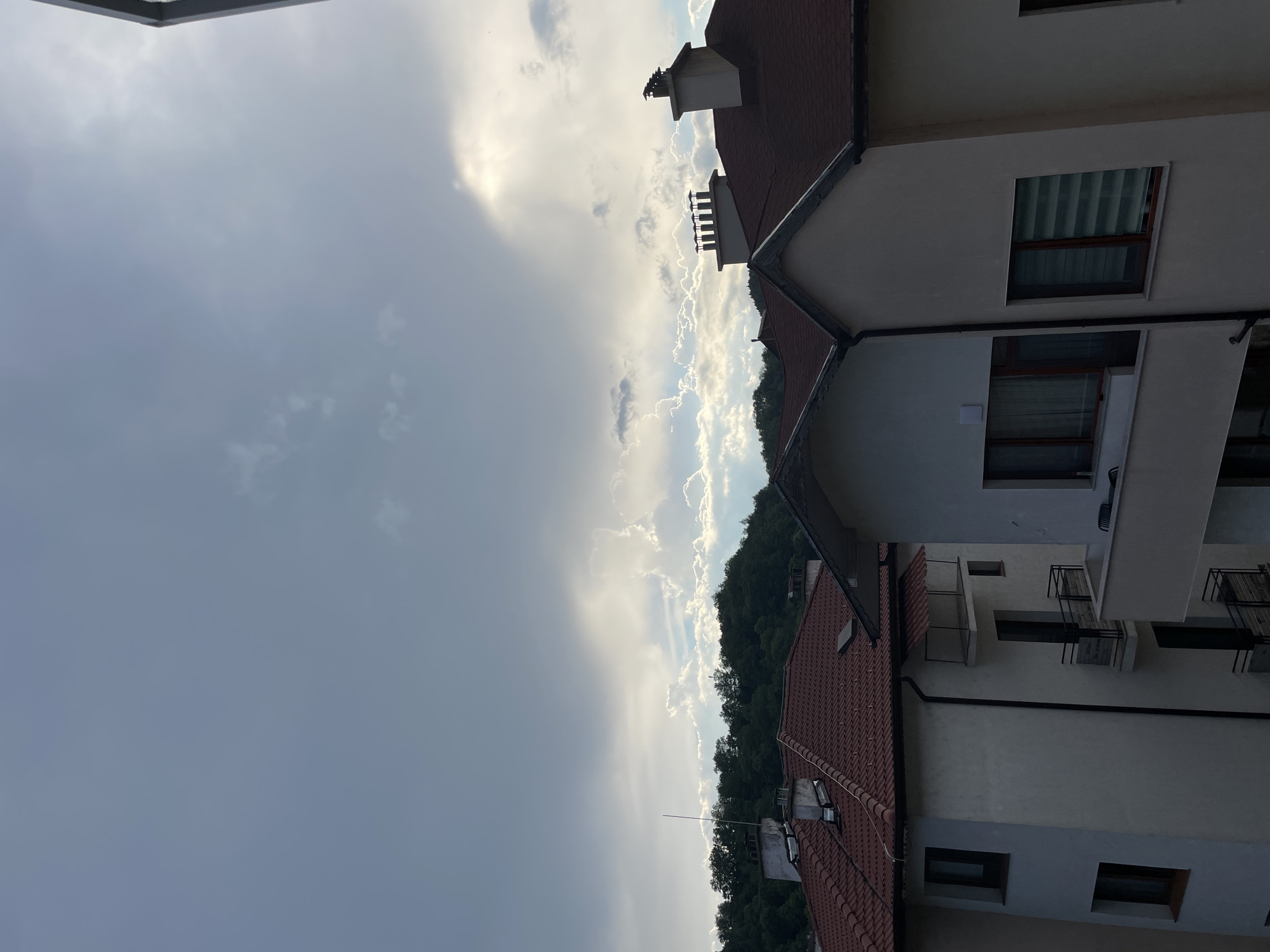 This is a photograph of a residential building with red tiled roofs in Zlatograd, Bulgaria. The image is oriented vertically, with the sky filling the left half and the building and a treeline on the right. The sky is dramatic, with a mix of dark storm clouds and bright, sunlit clouds, suggesting an approaching or receding storm. Sunlight breaks through the clouds in a bright, ethereal glow.
The building appears to be modern, with white walls and several windows. Some windows have blinds. Chimneys are visible on the rooftops, some with birds perched on them. There are also visible architectural features such as balconies with railings. A thin antenna is also visible on one of the roofs.
The treeline behind the building indicates a natural, possibly hilly or mountainous, landscape. The overall mood is one of awe at the power of nature, with the dramatic sky contrasting with the ordered structures of the buildings. The time of day is likely late afternoon or early evening, given the quality of the light. There is no visible text in the image.