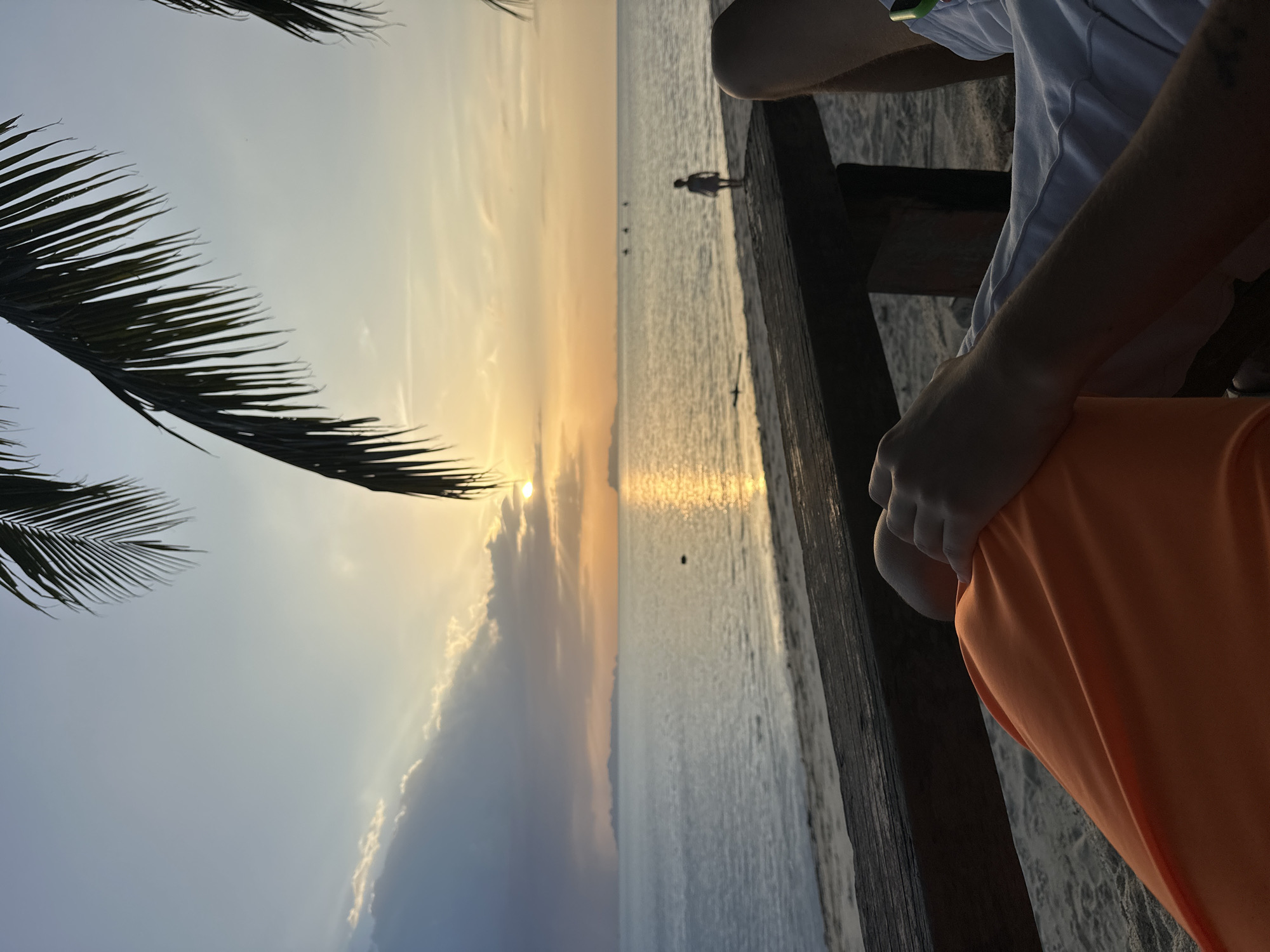 The image captures a serene sunset scene at a beach in Ko Samui District, Thailand. In the foreground, a person's legs, one clad in bright orange fabric and the other in white shorts, are visible, resting on a sandy surface. They appear to be seated on a wooden structure that stretches across the lower part of the frame. The middle ground features a wooden deck or pier running parallel to the water's edge. Beyond this, a calm sea reflects the warm hues of the setting sun, which is partially obscured by clouds, casting golden rays across the water. A solitary figure, appearing to be a child, stands on the sandy shore near the water, looking out towards the sea. In the distance, a faint silhouette of islands can be seen on the horizon, and several small boats are dotted across the water. Palm fronds frame the top of the image, adding a tropical touch to the tranquil vista.