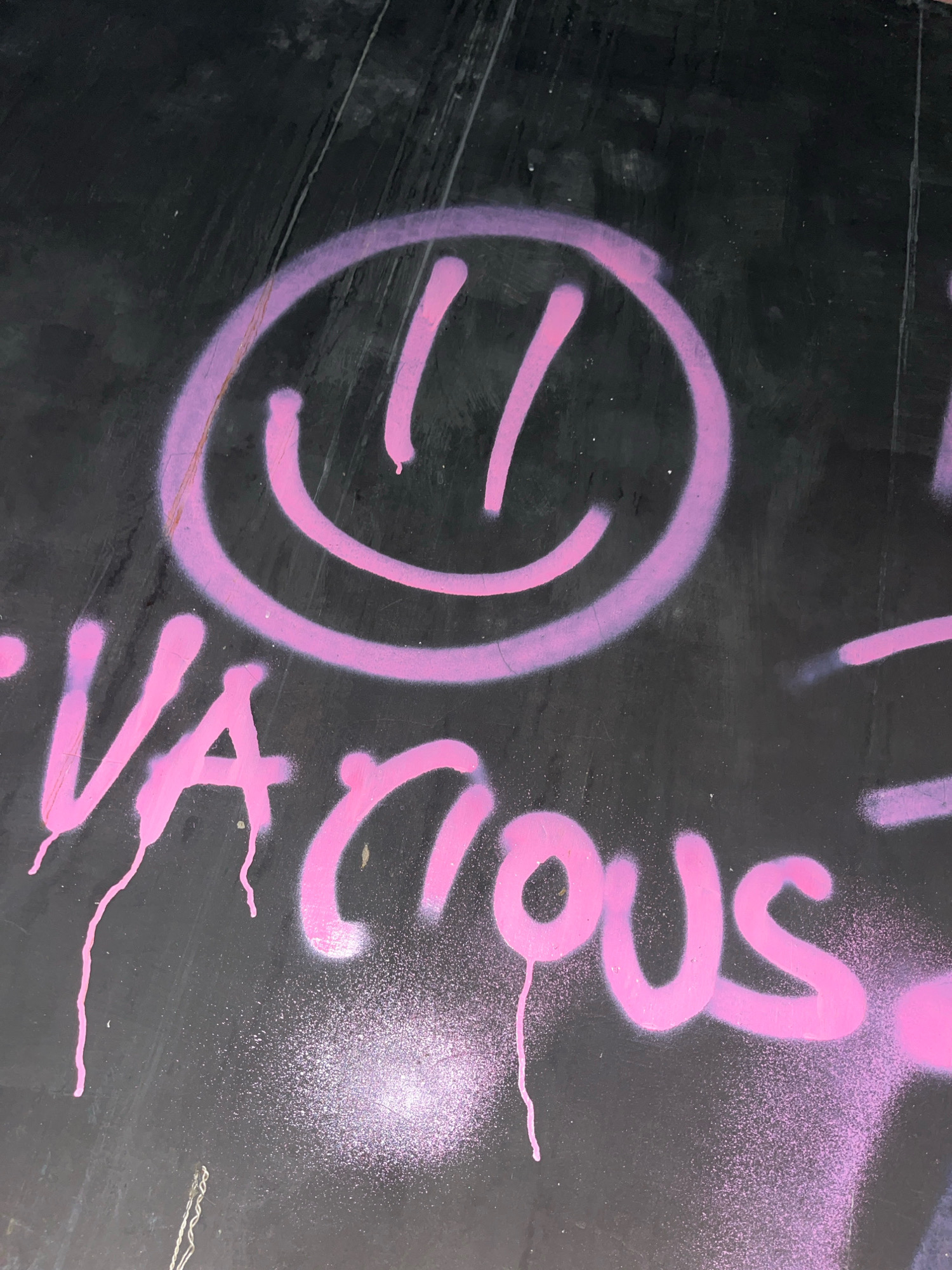 A close-up, low-angle shot captures graffiti on a dark, possibly metallic surface. The graffiti features a large pink smiley face in the upper half, with two vertical lines for eyes and a curved line for a mouth, all rendered with spray paint. Below the smiley face, the word "VARIOUS" is spray-painted in pink, with a drippy effect. The background is mostly black, with some visible lines and textures on the surface, suggesting it might be a wall or a vehicle. The lighting is dim, with a slight purple hue reflecting off the graffiti, possibly from the spray paint itself or ambient lighting conditions. The image appears to be taken in an urban environment in Surakarta, Indonesia, based on the provided context.
