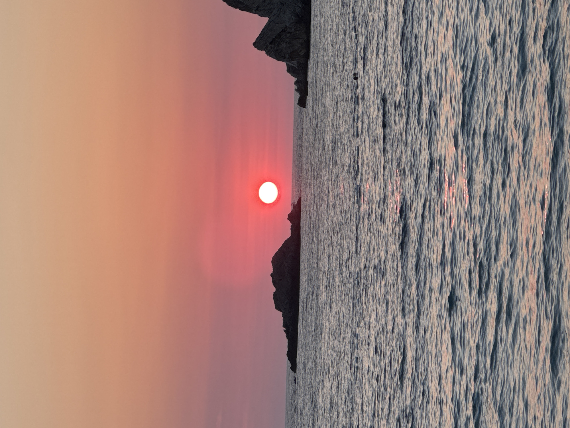 A vertical, close-up shot captures a vibrant sunset over the Adriatic Sea at Perazića Do, Montenegro. The sun, a brilliant orb of red and pink, hovers just above the horizon, casting a warm glow across the sky and reflecting on the rippling water. Jagged rock formations emerge from the sea on either side of the sun, silhouetted against the colorful sky. The water is dark blue with streaks of pink and orange from the sunset. In the middle distance, two dark figures are visible swimming in the sea. The overall impression is one of a serene and picturesque coastal scene.