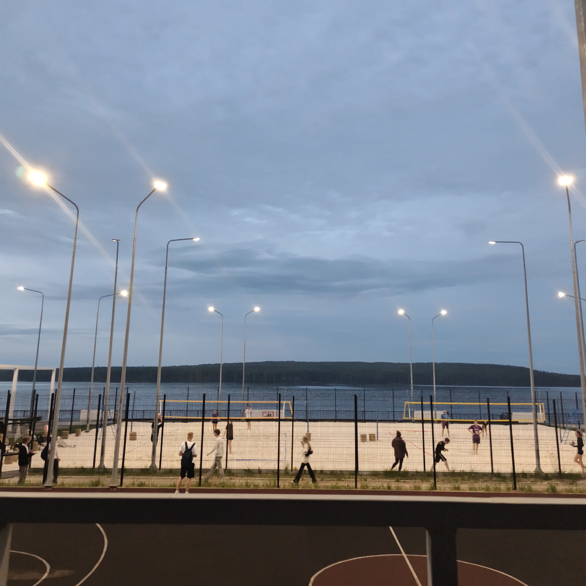 This media file captures an outdoor scene at dusk, likely in Chaikovsky, Russia. The foreground shows a dark, hard-surfaced court, possibly for basketball or another sport, with faint white markings. Beyond this, a fenced-off sand volleyball court is visible. Several people are present on the volleyball court, with some appearing to be playing a game and others observing or walking around.
The setting is a waterfront area, with a body of water stretching across the midground, bordered by a dark, forested treeline in the distance. Numerous tall streetlights are positioned along the waterfront, illuminating the scene as daylight fades. The sky is overcast with muted blues and grays, suggesting the time is either early evening or predawn. The overall mood is calm and recreational. No text is visible in the image.