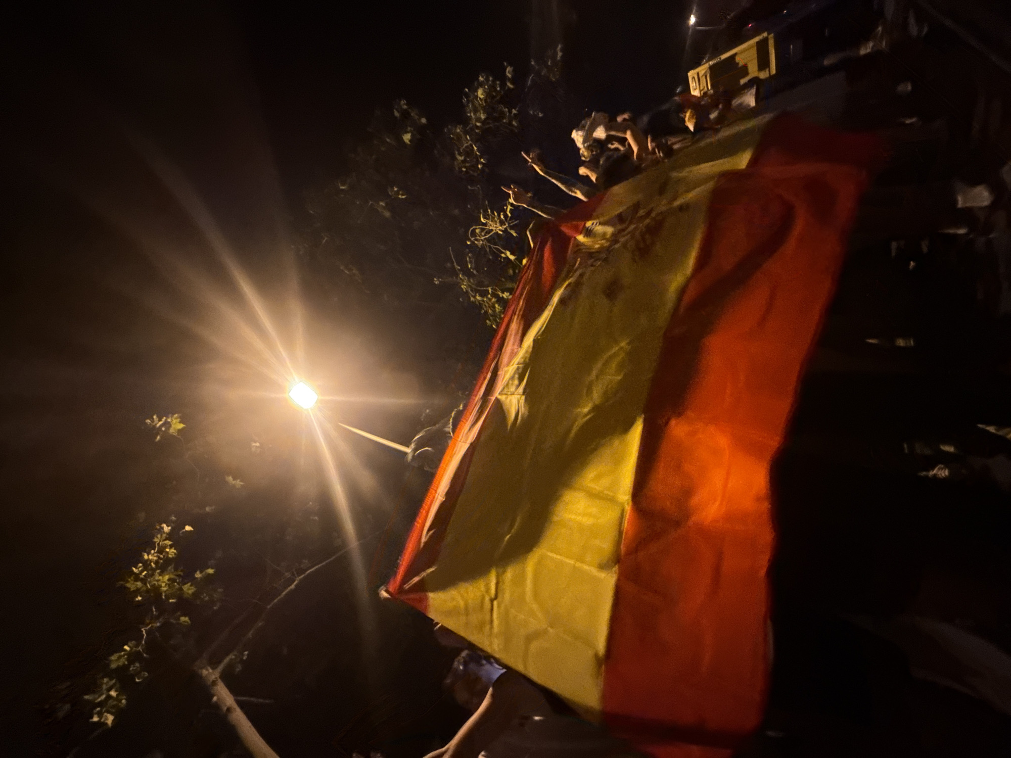 The image captures a nighttime street scene in Barcelona, Spain, illuminated by a bright streetlamp. A large Spanish flag, predominantly red and yellow, is prominently displayed, held aloft by several people whose faces are largely obscured by the flag and the darkness. The flag appears to be a focal point of the gathering, suggesting a celebratory or patriotic event. In the background, trees with sparse foliage are visible against the dark sky, and the light from the streetlamp creates lens flares and a diffused glow. Other individuals are present, with some hands raised in the air, possibly in excitement or support. The overall atmosphere appears energetic and communal, centered around the display of the national flag. The presence of what appears to be a yellow structure, possibly a vehicle or temporary setup, is visible on the right side of the frame.