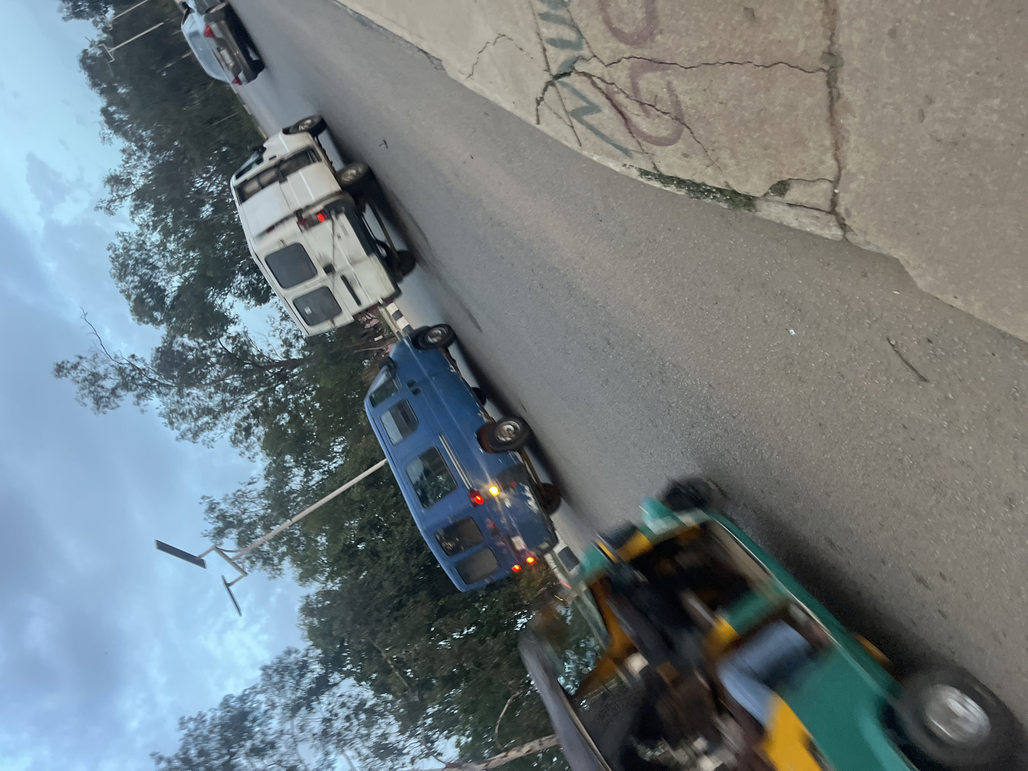 In Gura Top, Nigeria, a street scene unfolds under an overcast sky, depicted with a slight motion blur. The perspective is from a low angle, looking across a paved road lined with trees. In the foreground, a three-wheeled auto-rickshaw, painted in teal and yellow, is in motion, with a person visible in the driver's seat wearing blue jeans.
Further down the road, a blue minibus is traveling, its brake lights illuminated. To its right, a white panel van with its rear door ajar is also in motion. Behind these vehicles, a gray sedan is visible on the road, and more trees line the background. A street light with two solar panels is positioned to the left of the frame, and the concrete curb on the right side of the road shows cracks and graffiti. The overall impression is of a typical street in a Nigerian city during daylight hours.